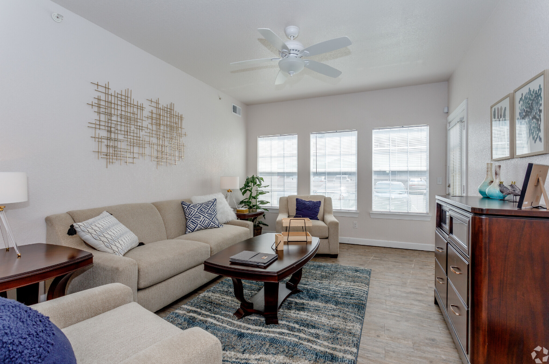 Cozy living room with tan sofa, blue rug, wooden furniture, and bright windows.