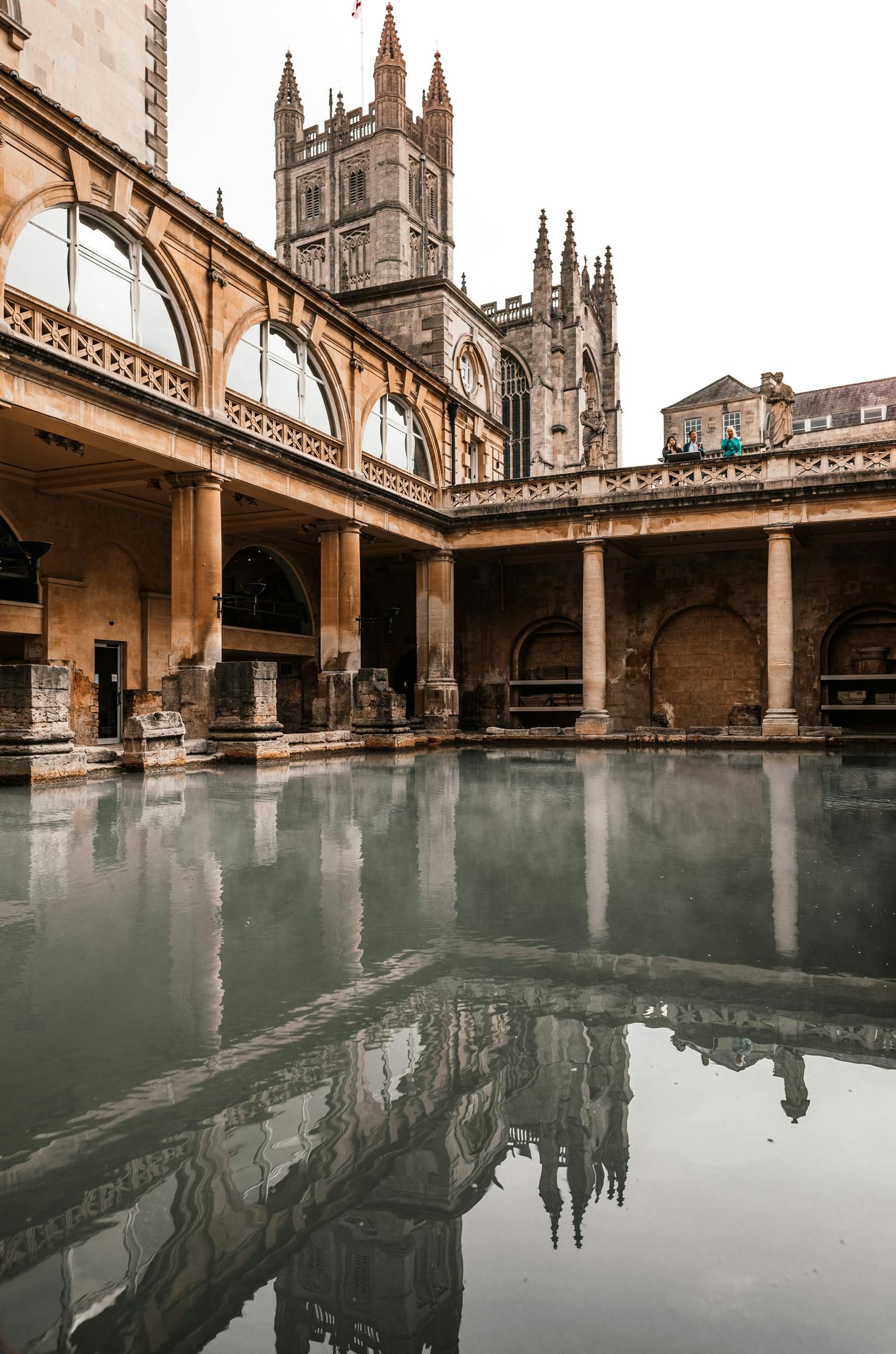 The Roman Baths in Bath, England. Ancient stone pool reflecting surrounding structures and Gothic cathedral in background.