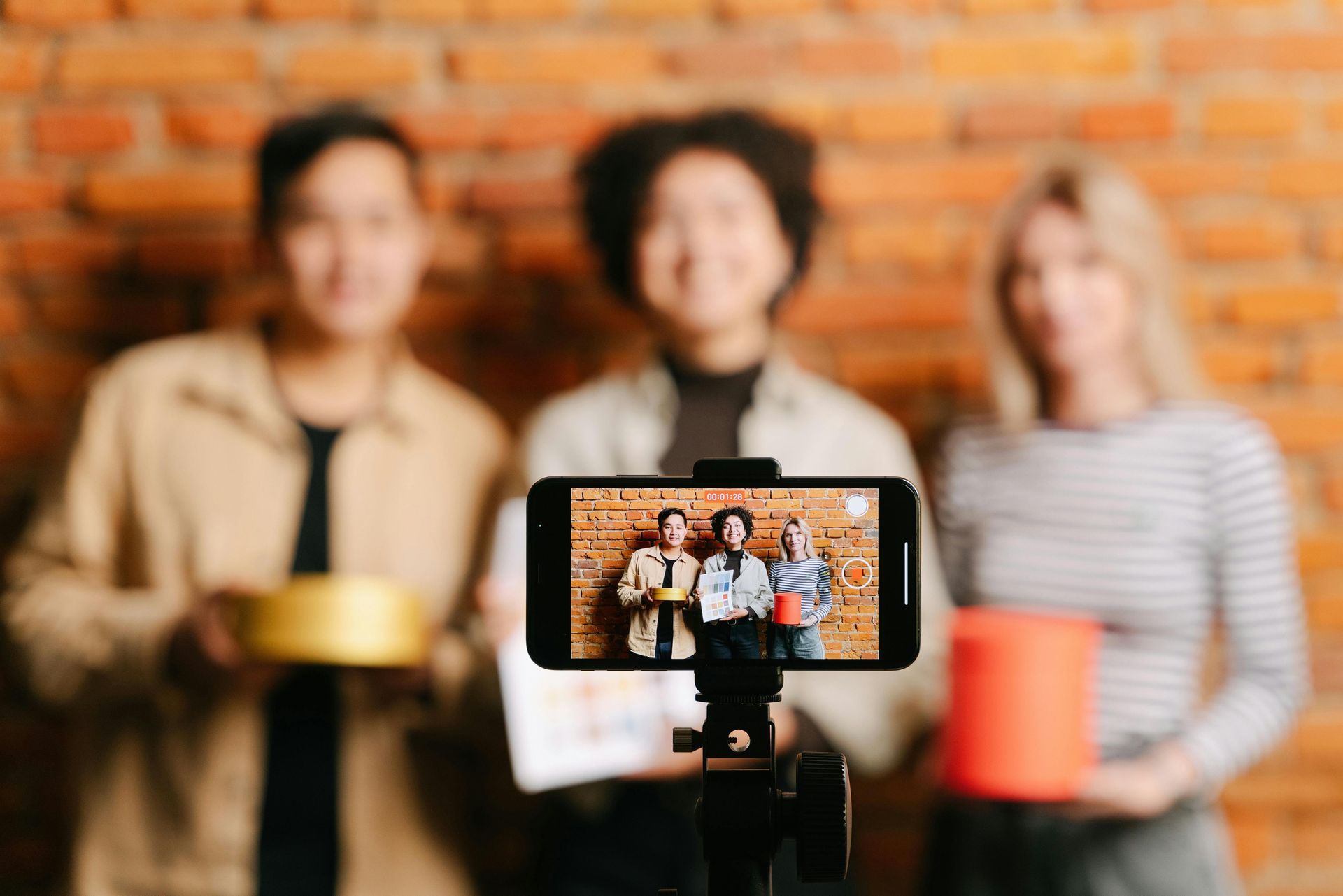 Three people filming a video in front of a brick wall, two holding objects, smiling.