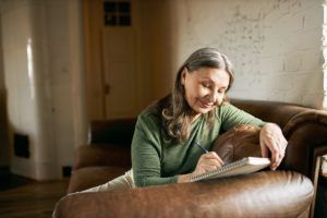 Creative beautiful middle aged woman with long gray hair sitting on comfortable leather couch with notebook handwriting poems, drawing or making list to do, having dreamy facial expression, smiling