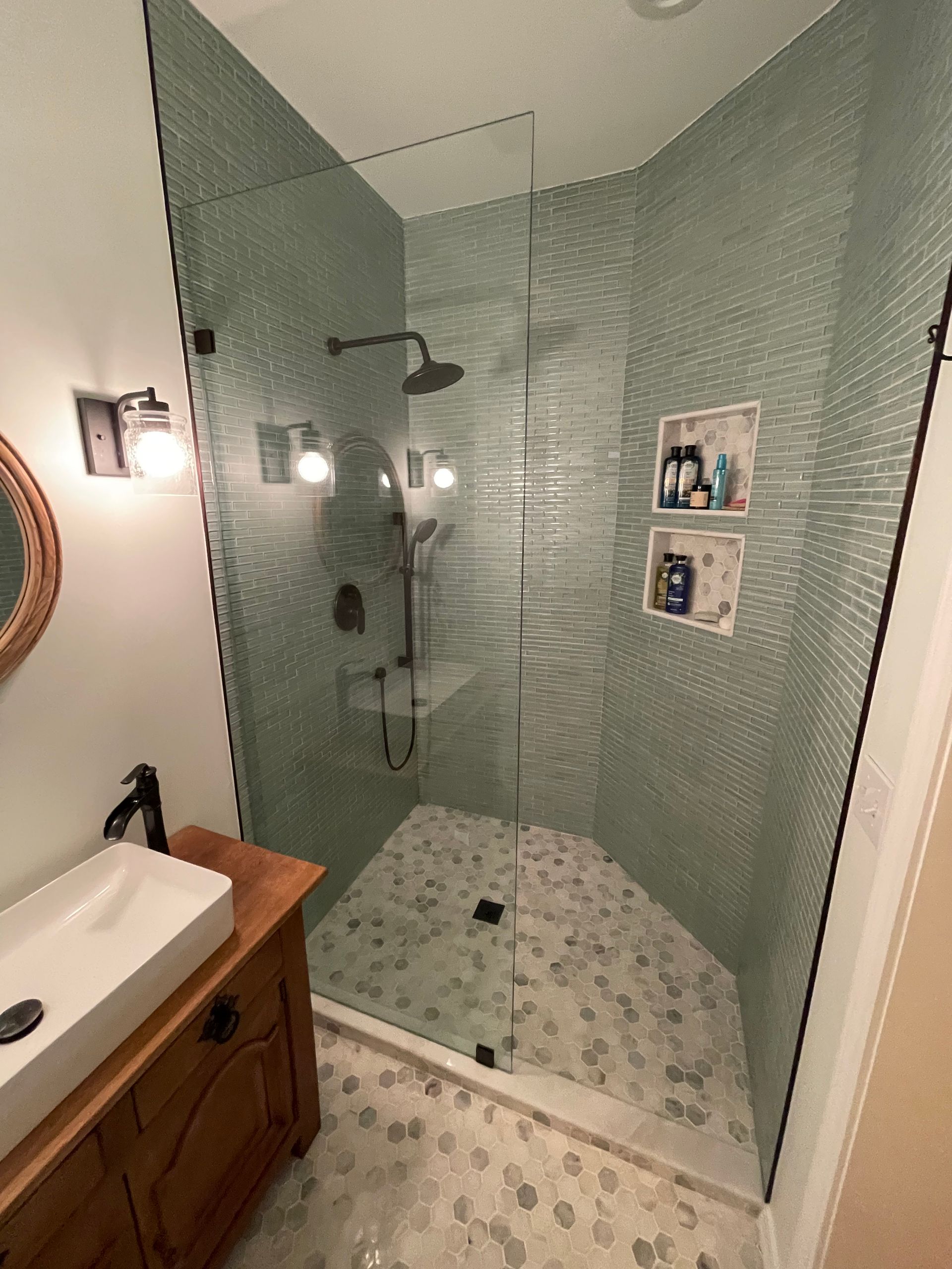 Modern bathroom with glass shower enclosure, green tiled walls, and stone floor.  Wooden vanity with vessel sink on the left.