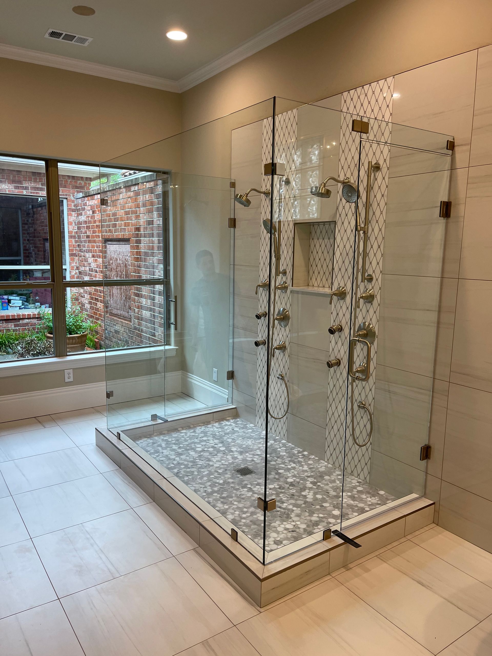 A large, glass-enclosed shower with multiple showerheads, a tiled floor, and a window.