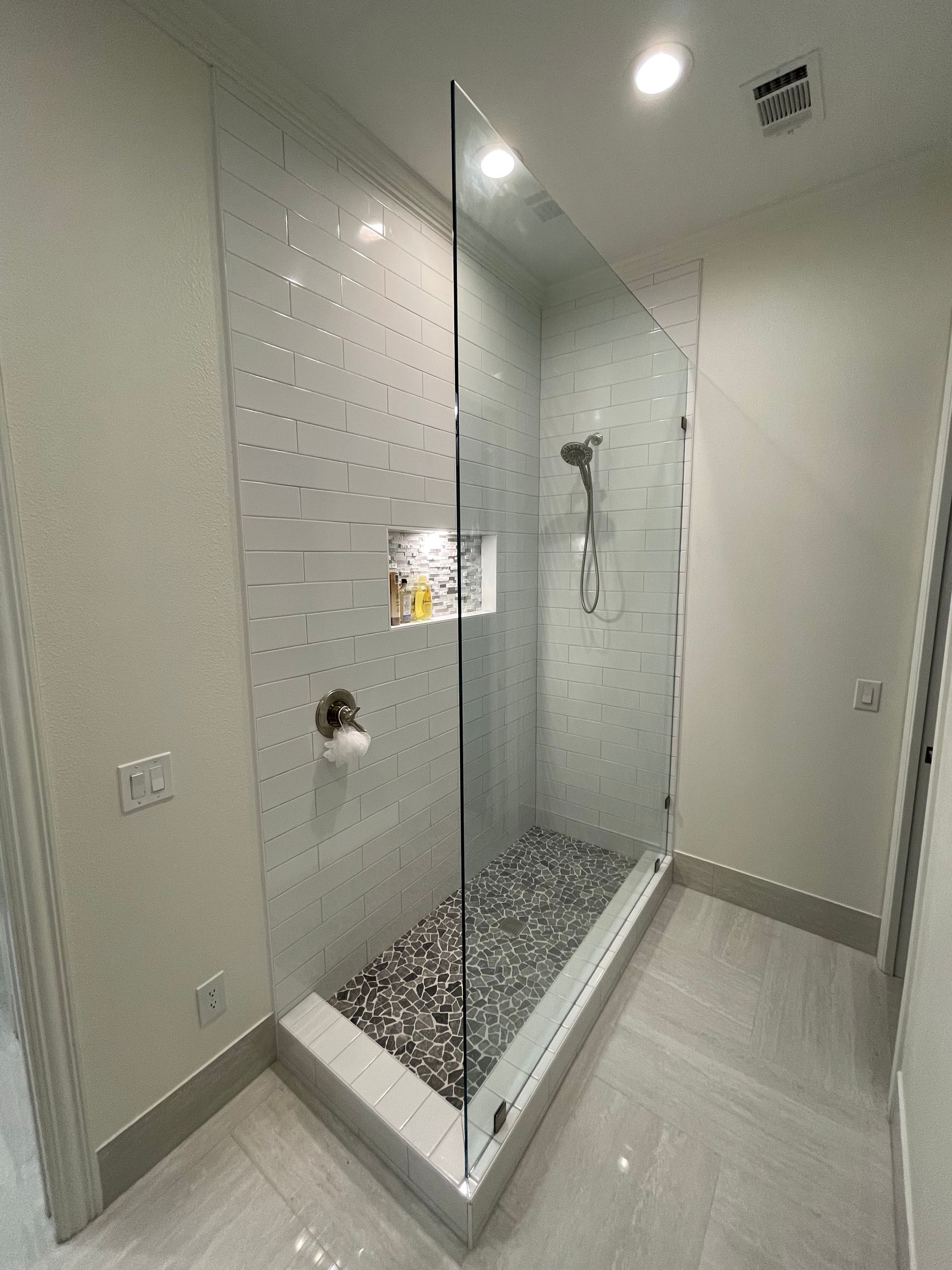 Glass-enclosed shower with pebble floor, gray tile walls, and recessed storage in a modern bathroom.