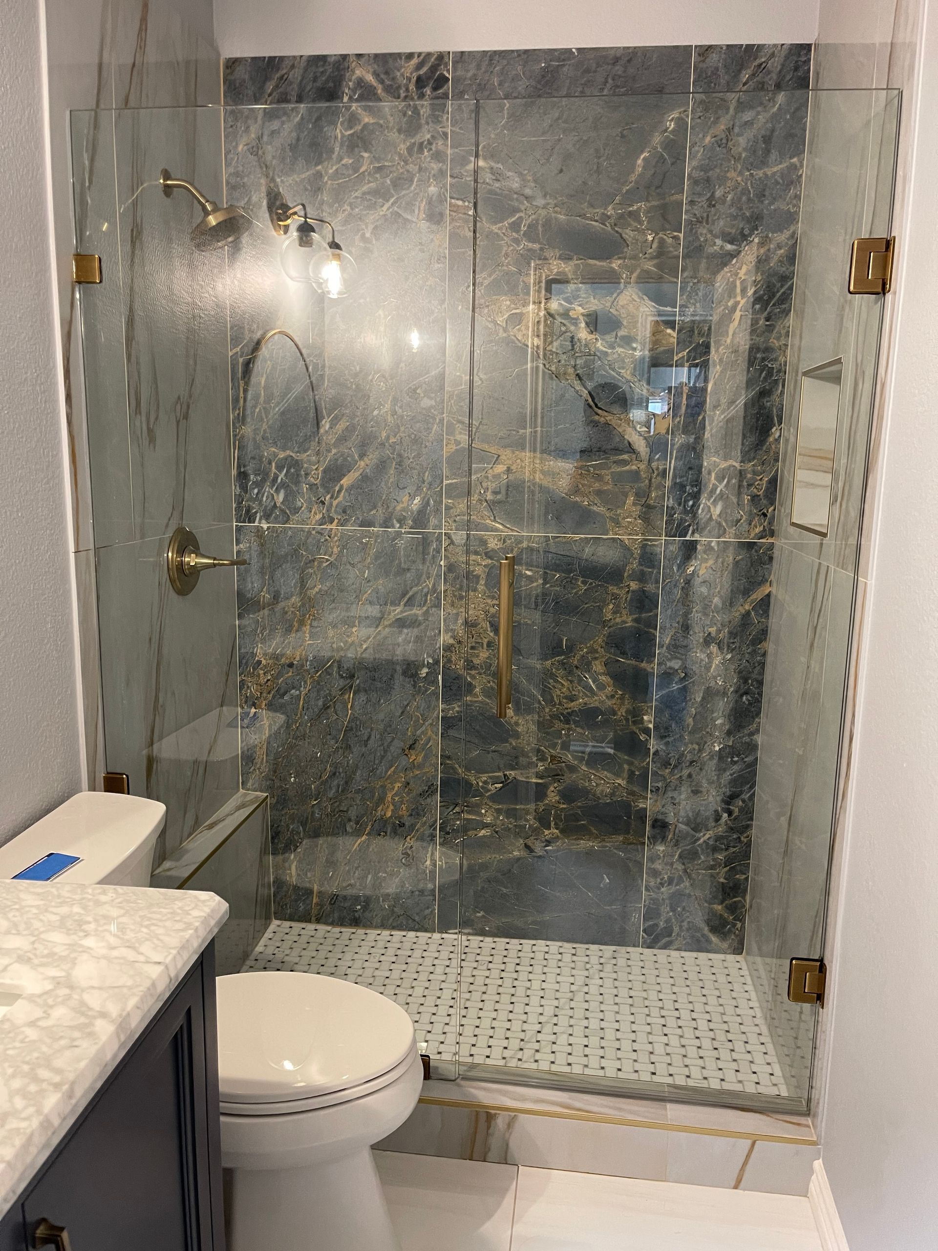 Bathroom with marble walls and a glass shower door; gold fixtures, blue vanity, and a white toilet.