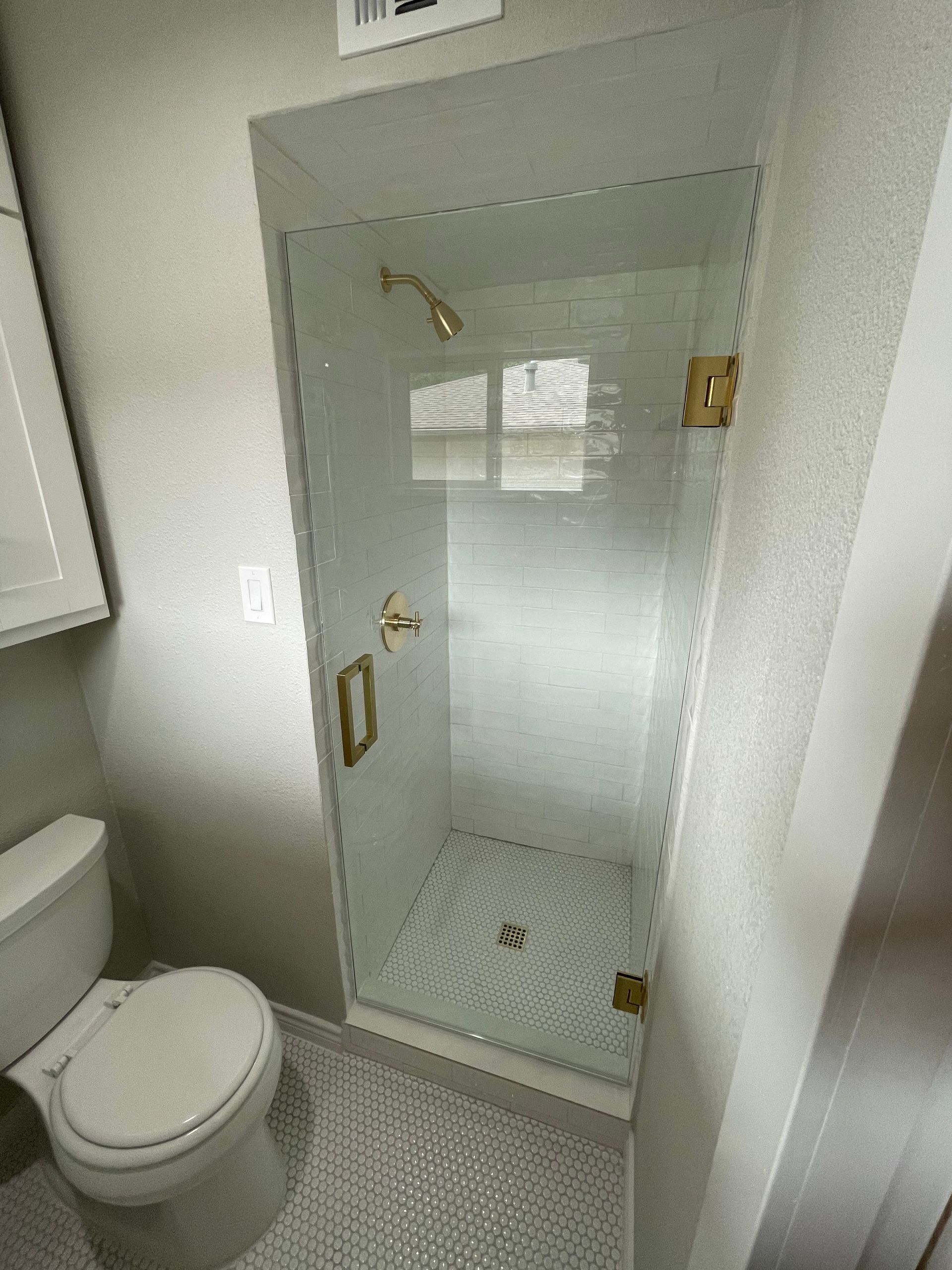 Small, modern shower with glass door and gold fixtures in a bathroom with white and gray tile.