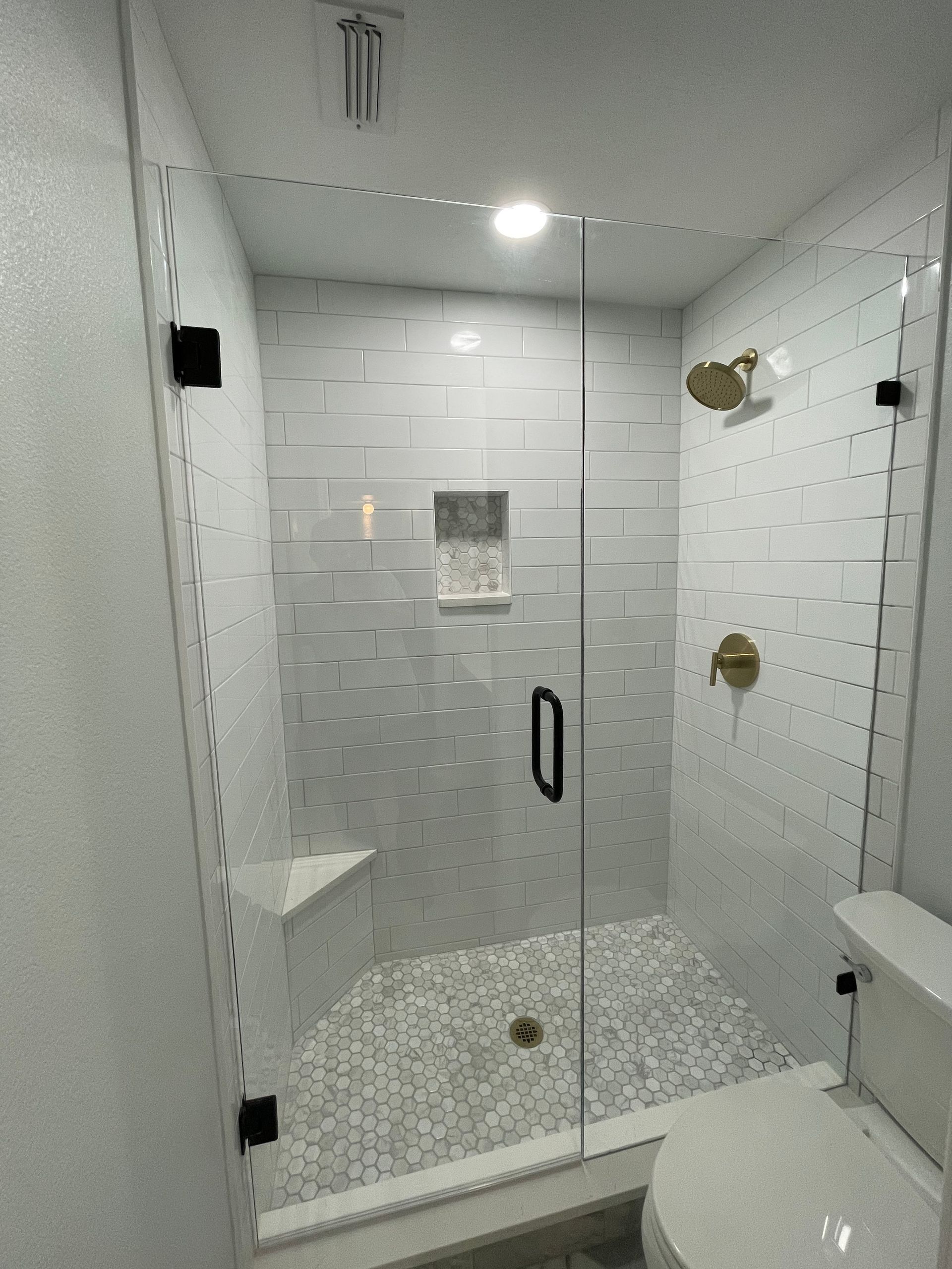 Modern bathroom with glass shower enclosure, white tile, gold fixtures, and pebble flooring.