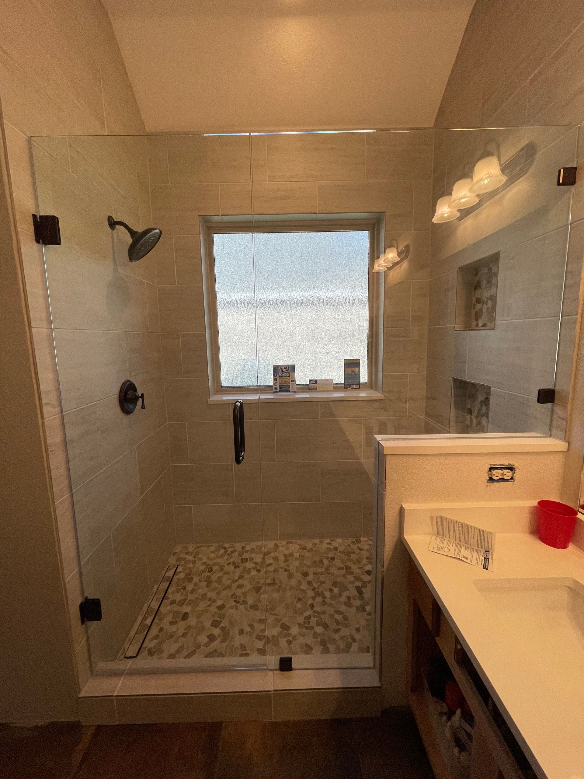 Shower with glass doors and pebble floor, beige tile walls. Window visible in back.