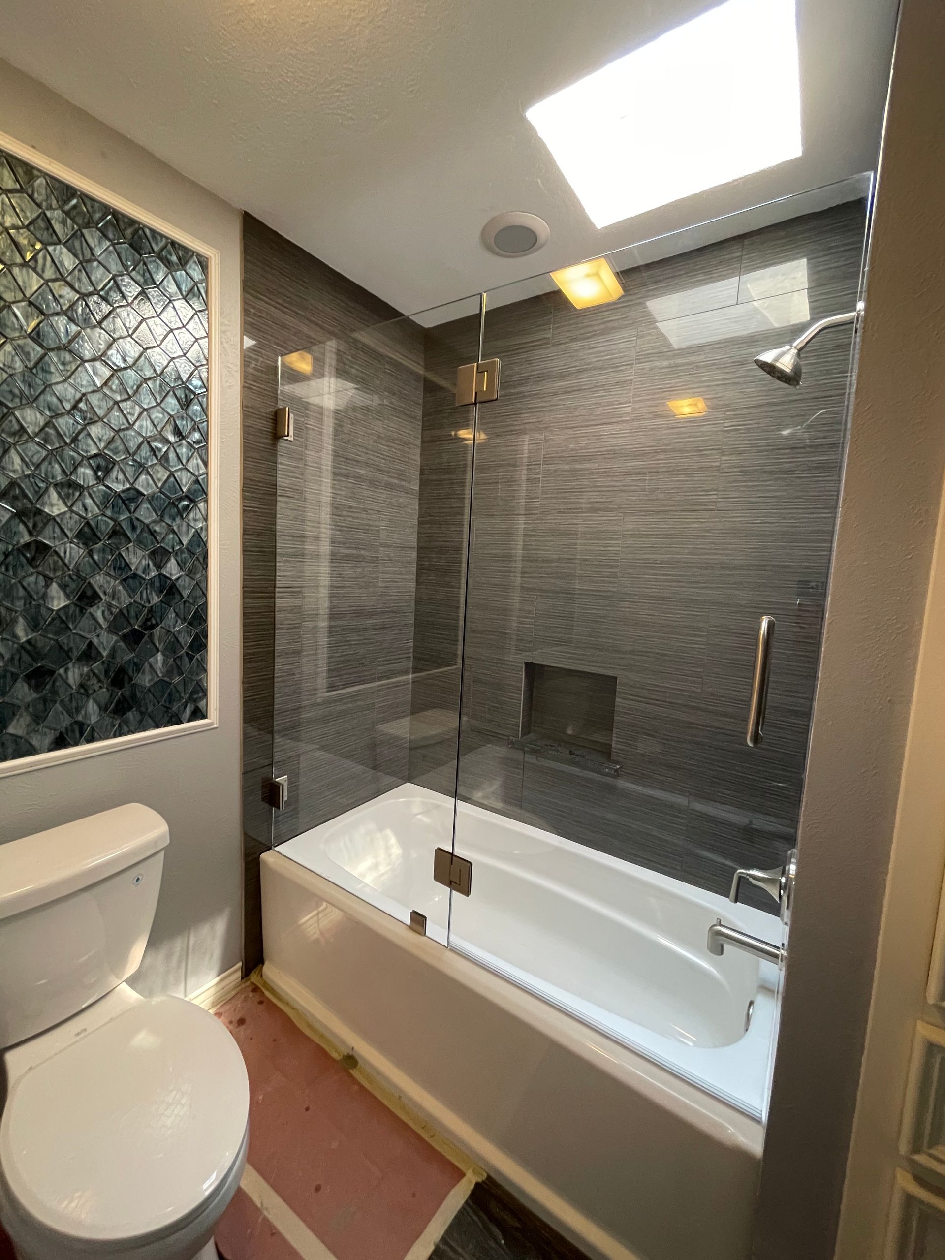 Bathroom with a white bathtub, gray tiled walls, glass shower enclosure, and toilet.