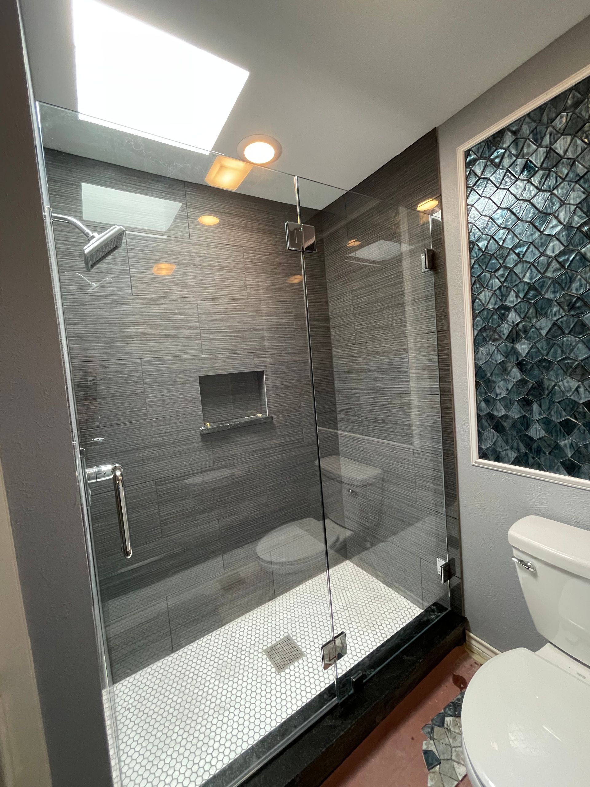 Modern bathroom with a glass-enclosed shower, dark tiled walls, skylight, and decorative tile accent wall.