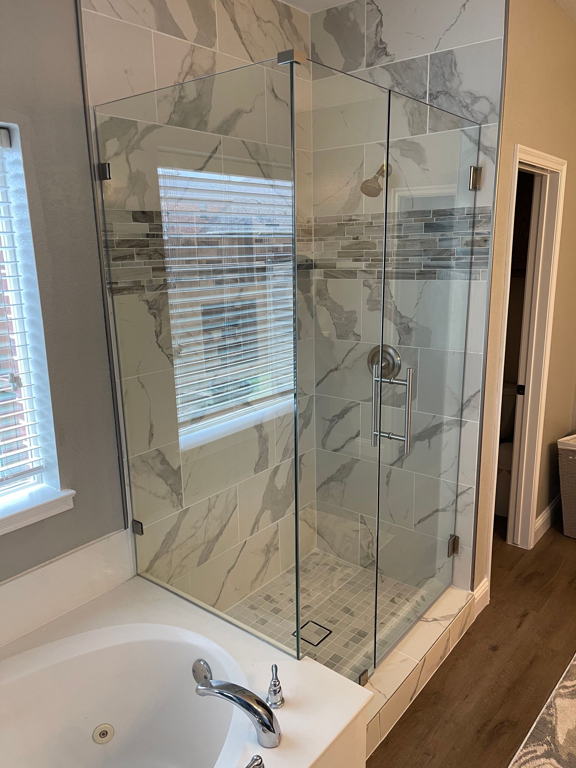 Glass shower enclosure with marble tile, chrome fixtures, next to a bathtub.
