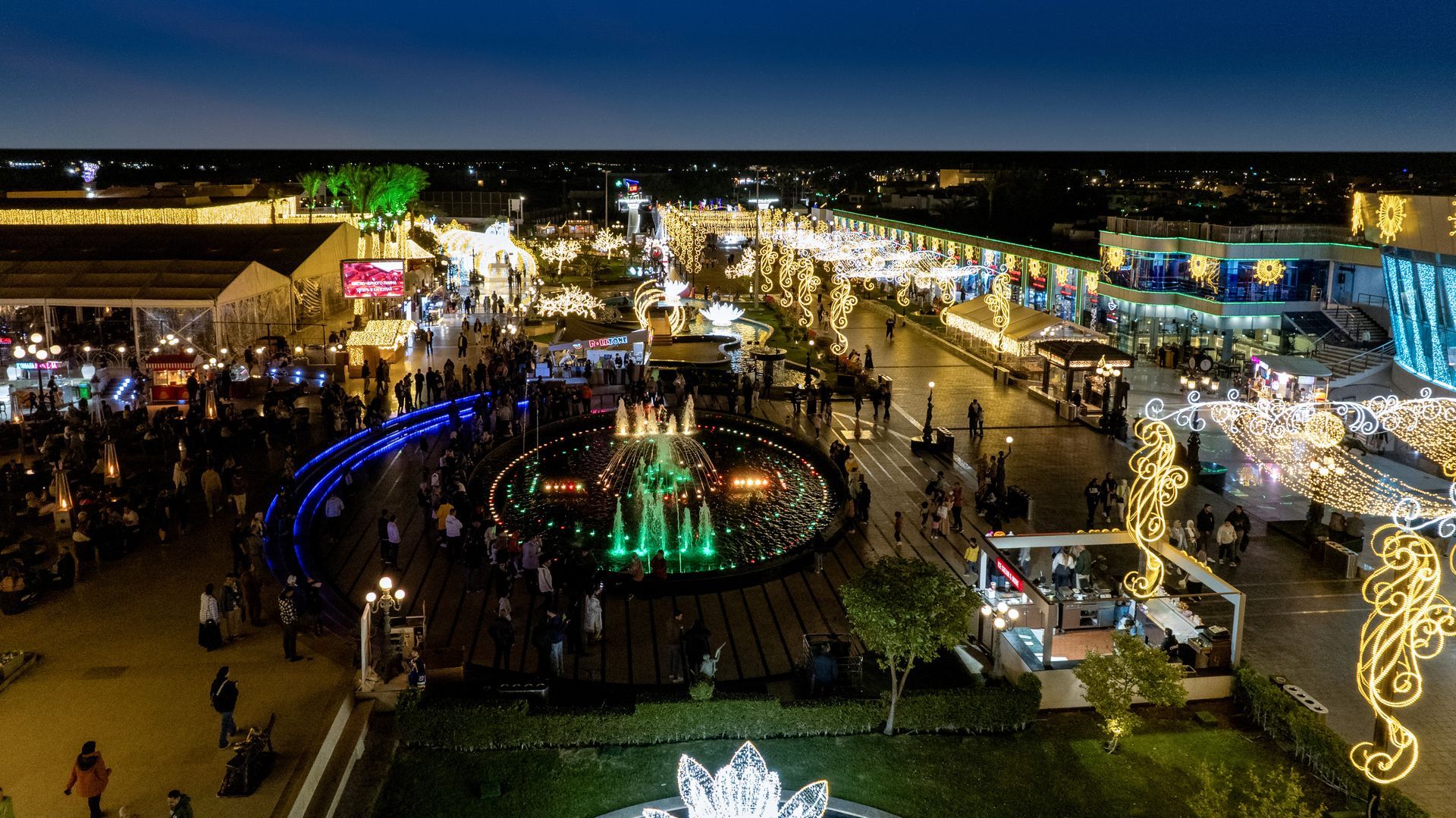 Nighttime aerial view of a lit-up outdoor plaza with people, shops, fountains, and festive lights.