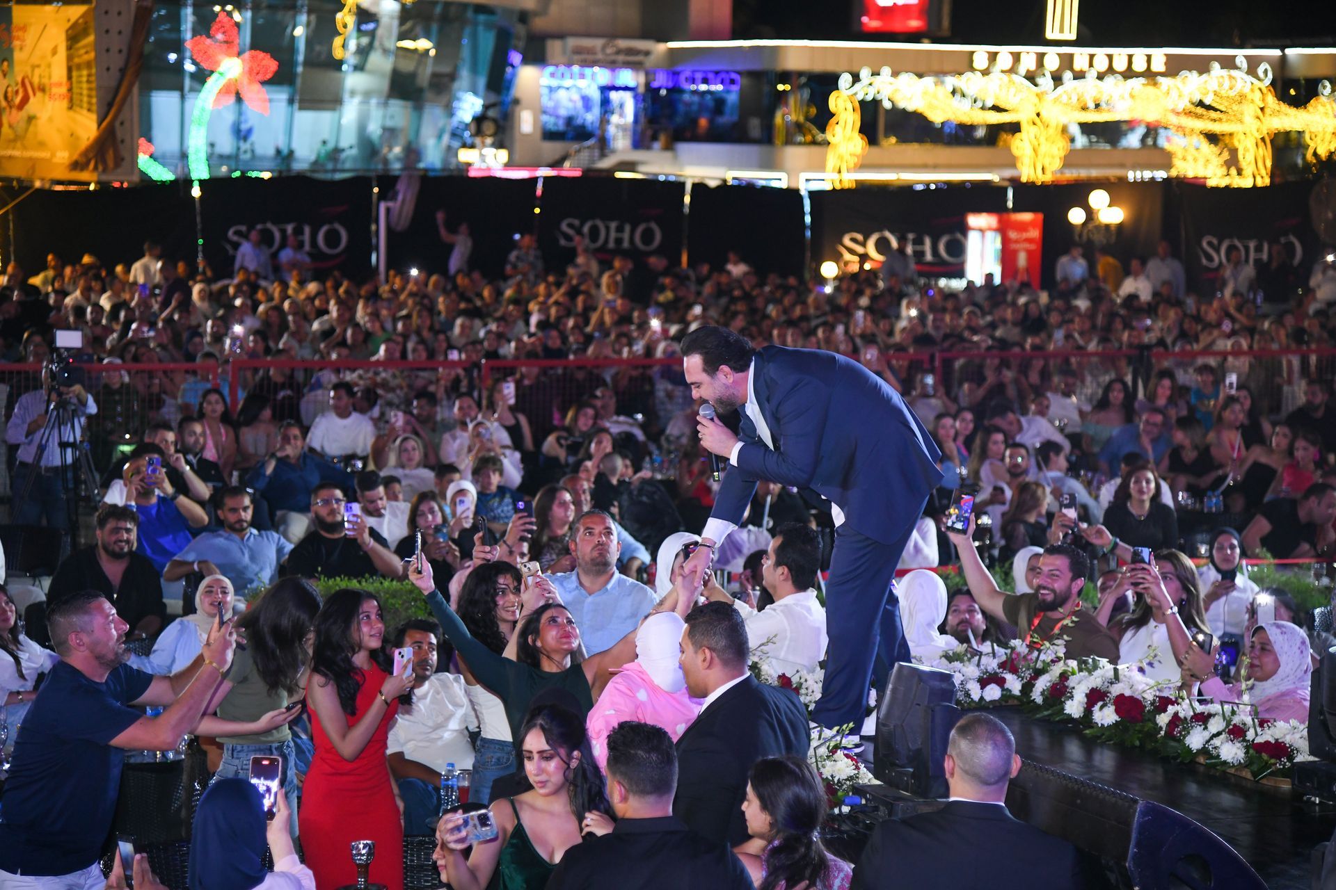 Man in suit sings on stage to a large, excited crowd at an outdoor event.