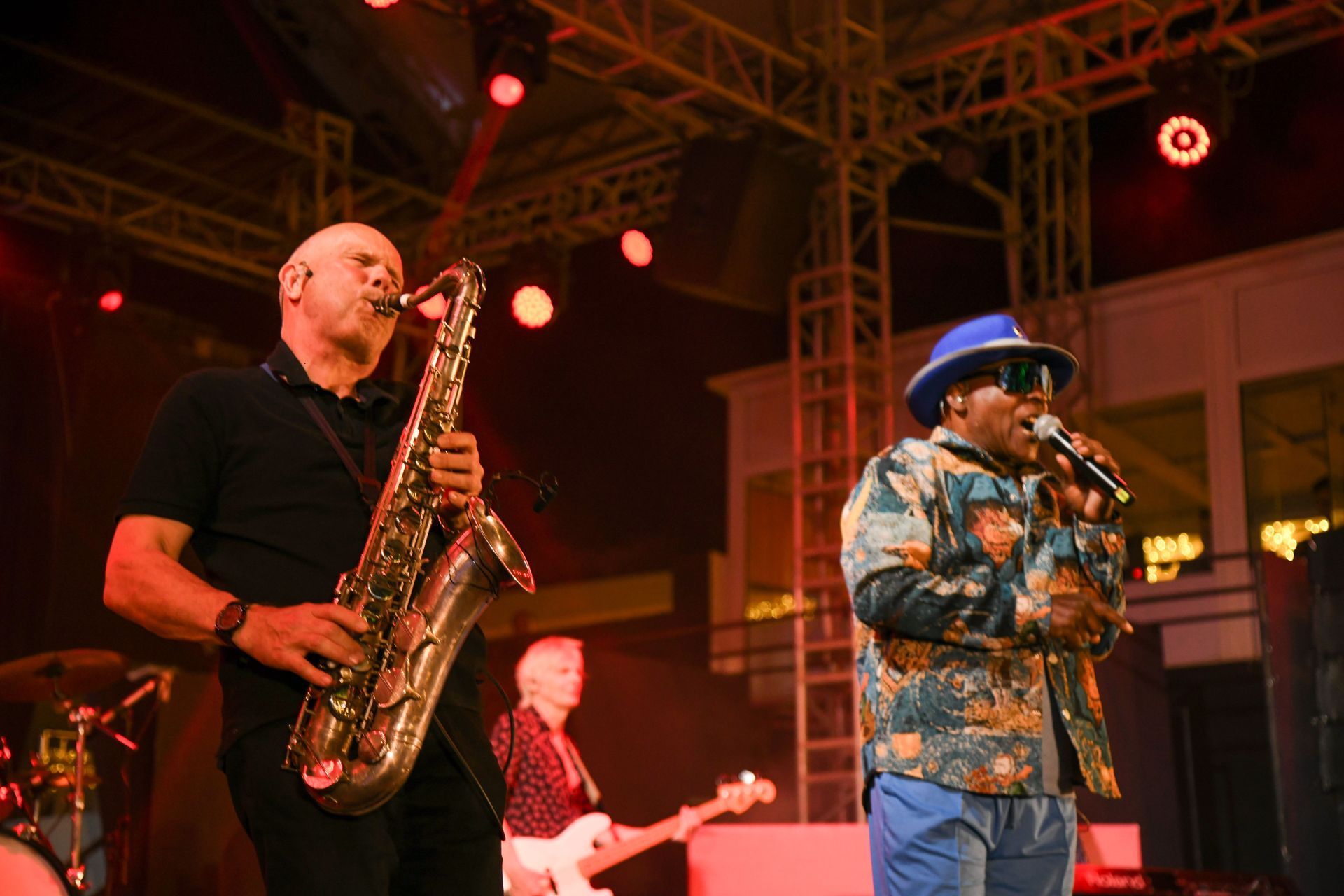 Saxophonist and singer on stage, performing with band. Red lights illuminate the scene.
