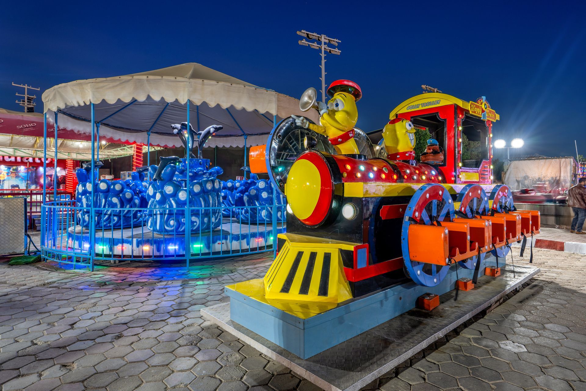 A colorful toy train and carousel illuminated at night in an outdoor carnival setting.