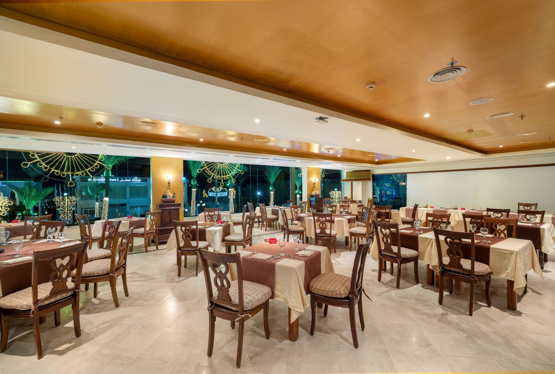 Restaurant interior with tables, chairs, and large windows overlooking outdoor lights at night.