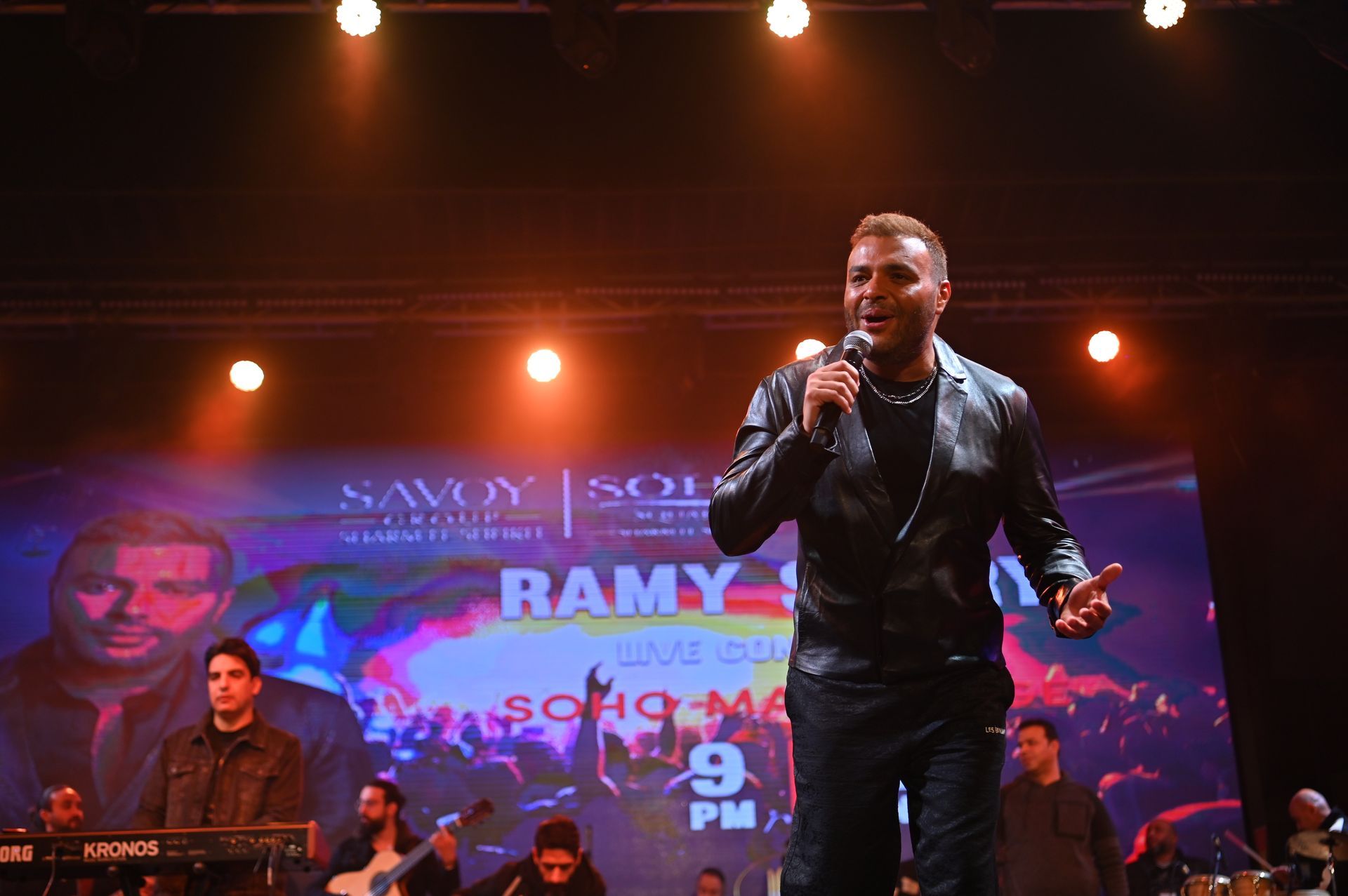 Man in black leather jacket sings on stage with a band; a banner advertises his name 