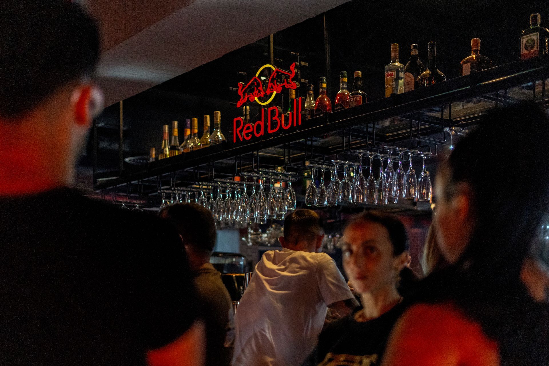 People at a bar, a Red Bull neon sign, bottles on shelves, and glasses hanging.