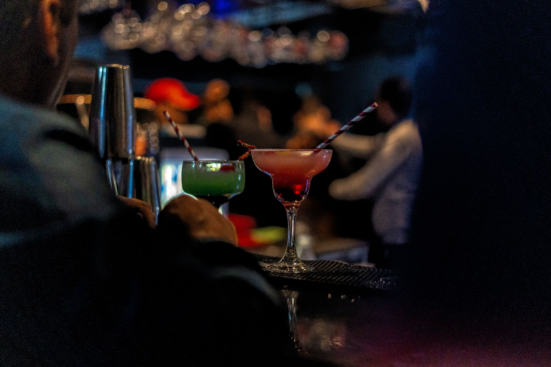Two cocktails on a tray at a bar. A bartender and patrons are in the blurry background.