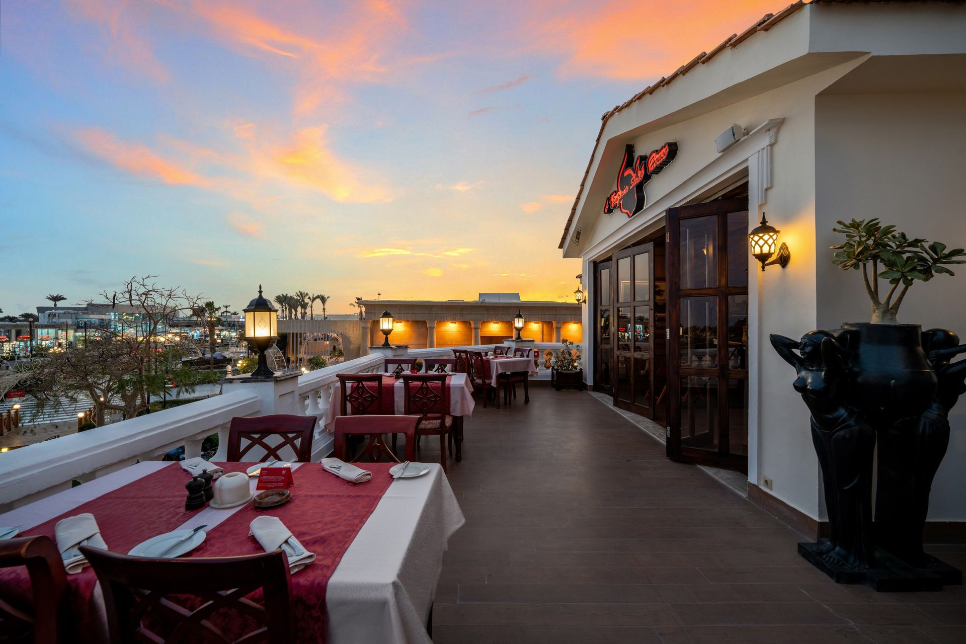 Rooftop restaurant at sunset. Tables with red tablecloths, wooden chairs. Views of buildings and a colorful sky.
