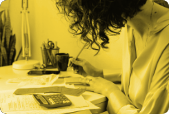 Woman working at a desk, using a pencil and calculator, lit in yellow.