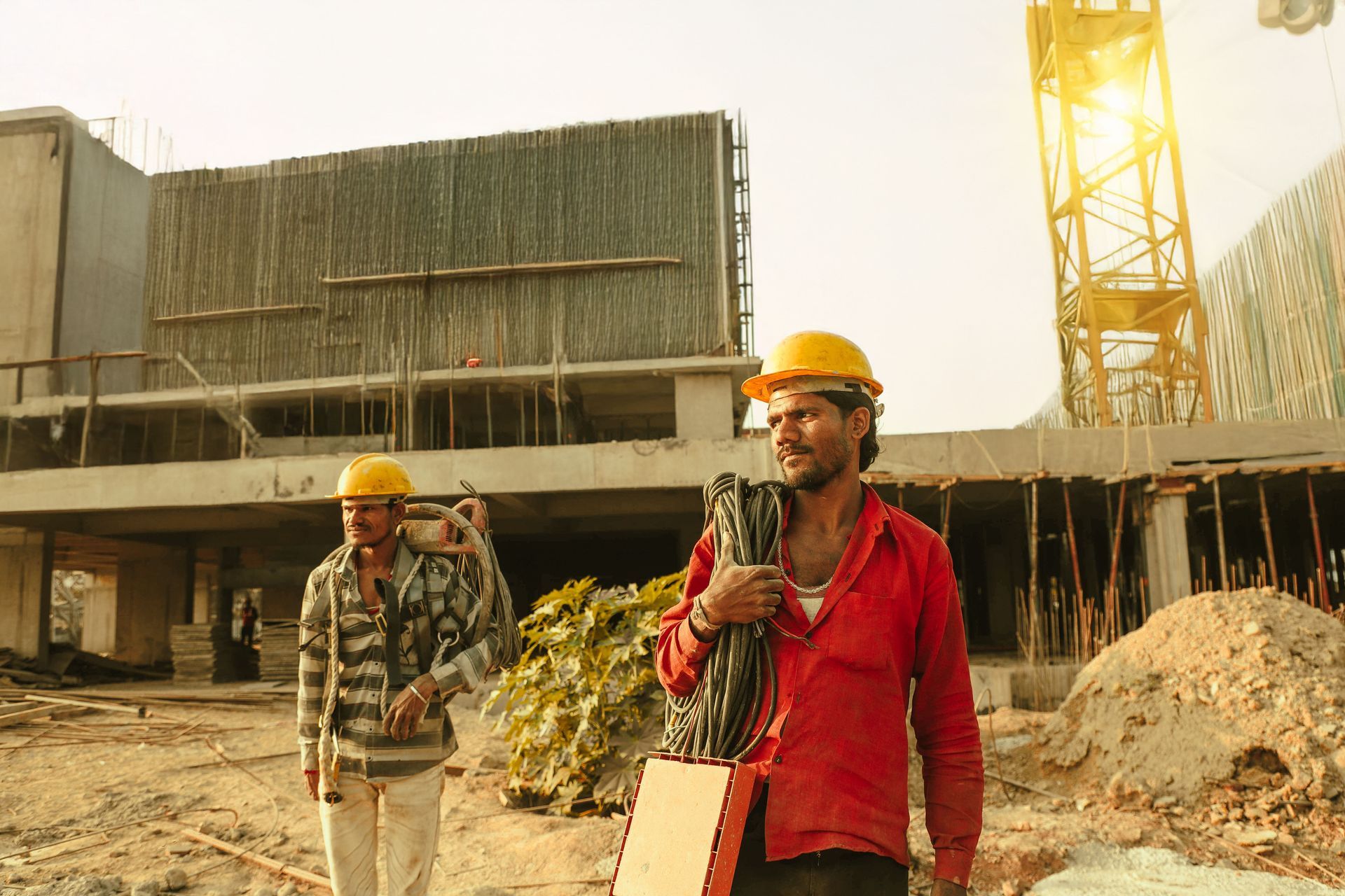 Two construction workers in hard hats walk at a dusty building site near a crane.