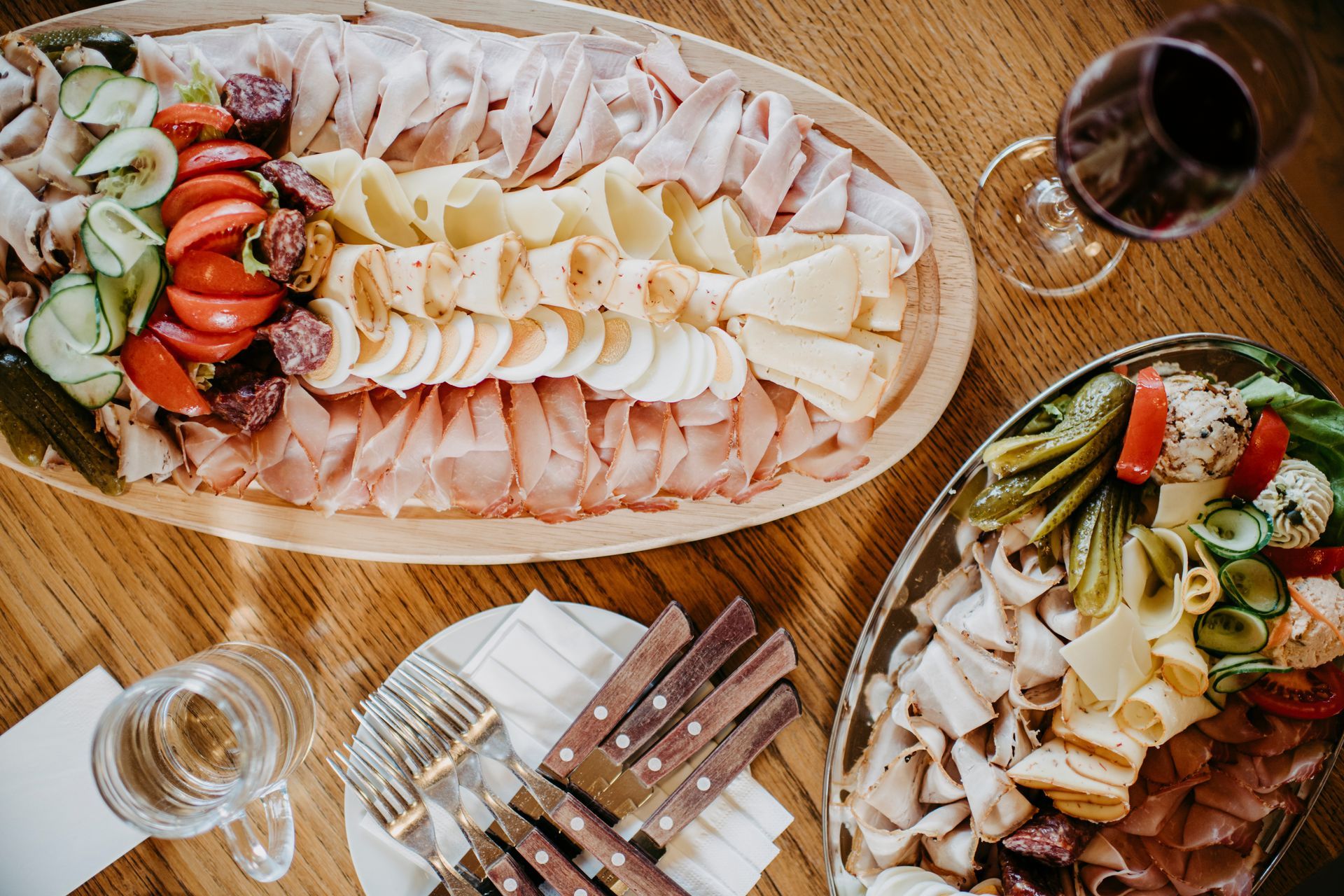 Wurst- und Käseplatten mit Fleisch, Käse und Gemüse, dazu ein Glas Wein und Besteck auf einem Holztisch.