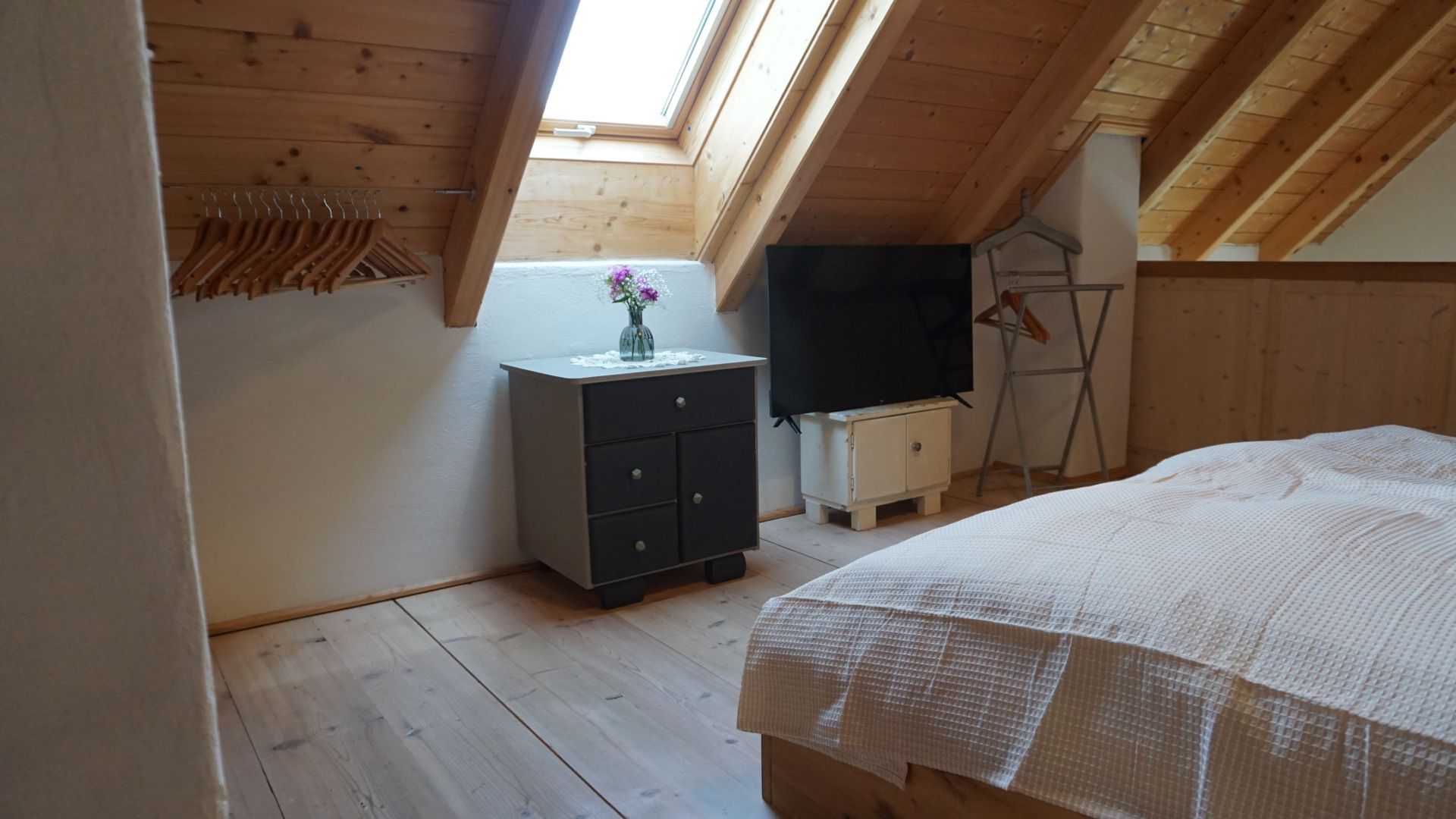 Schlafzimmer mit Holzdecke, Dachfenster, Bett, Fernseher, Schrank und Kleiderbügeln.