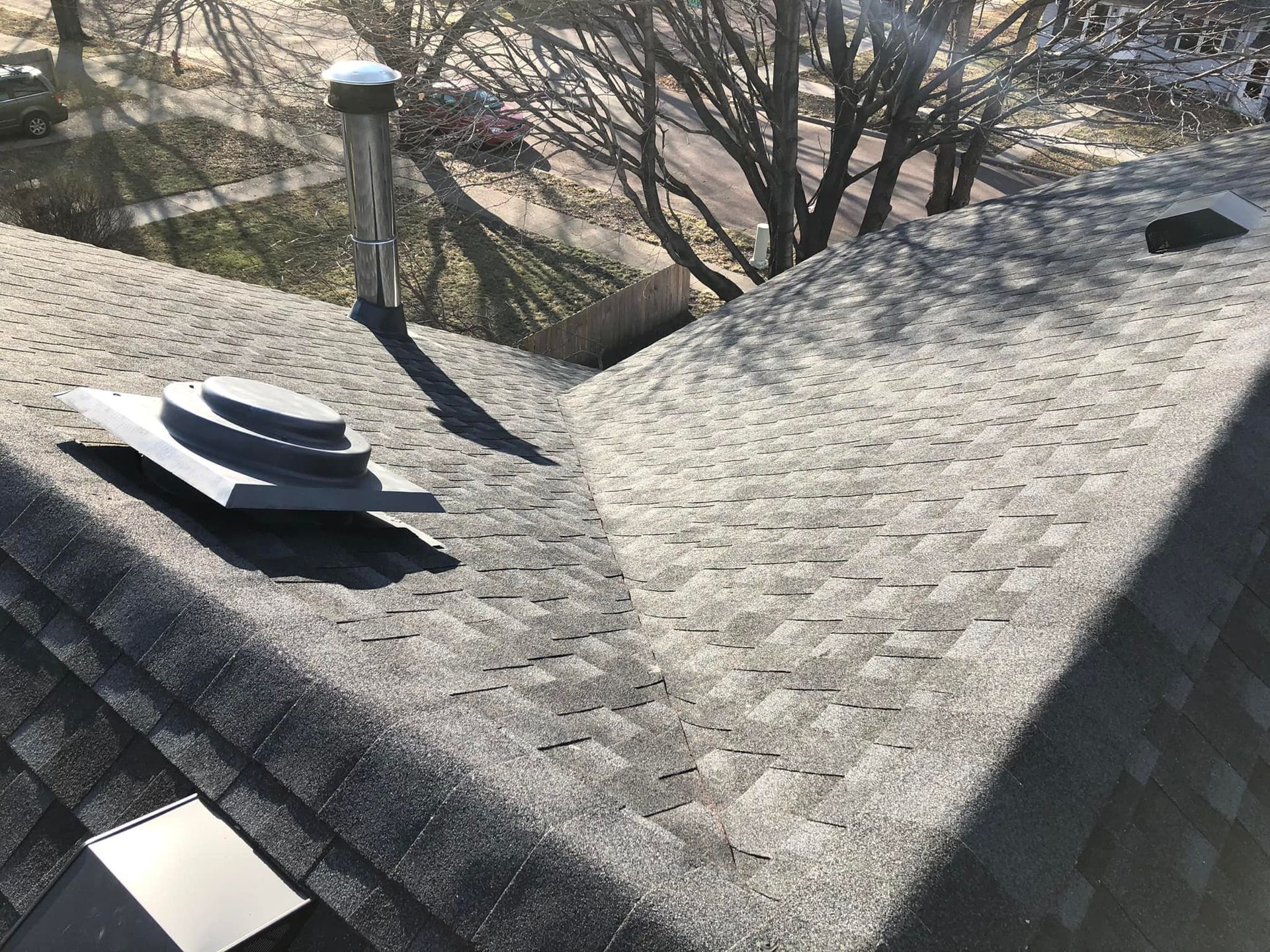A grey shingle roof with a silver chimney and a grey vent on a sunny day.