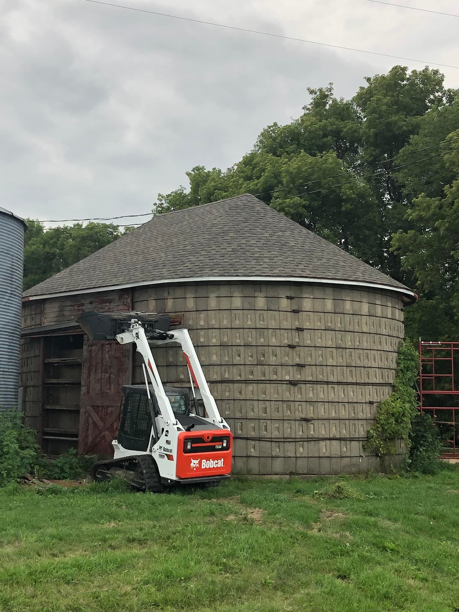A Bobcat skid-steer loader parked near a rustic silo on a grassy field. Overcast sky.