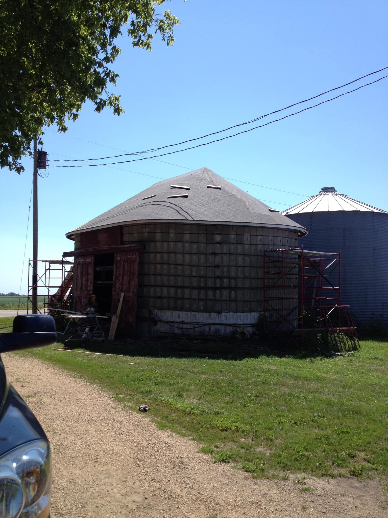 Old wooden round barn with a weathered roof, red doors, and a silo in the background on a sunny day.