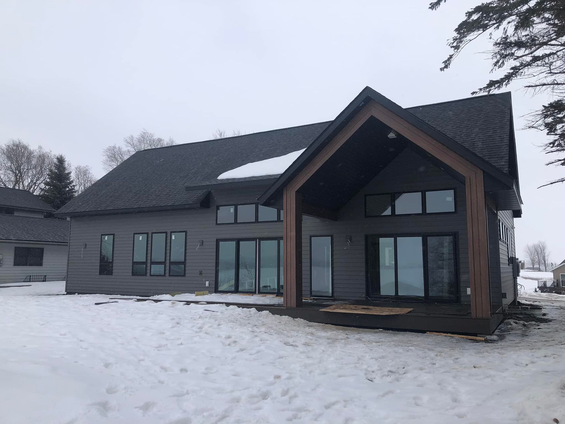 Modern gray house with black roof, windows, and snow-covered yard. Wooden porch, winter scene.