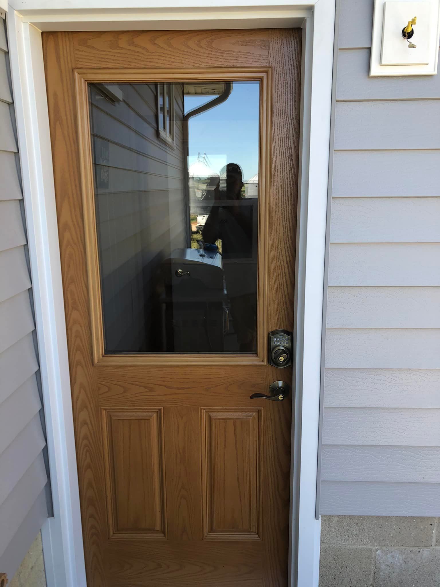 Wooden door with a glass panel, exterior view. Light brown door with a lock, surrounded by white trim and blue siding.