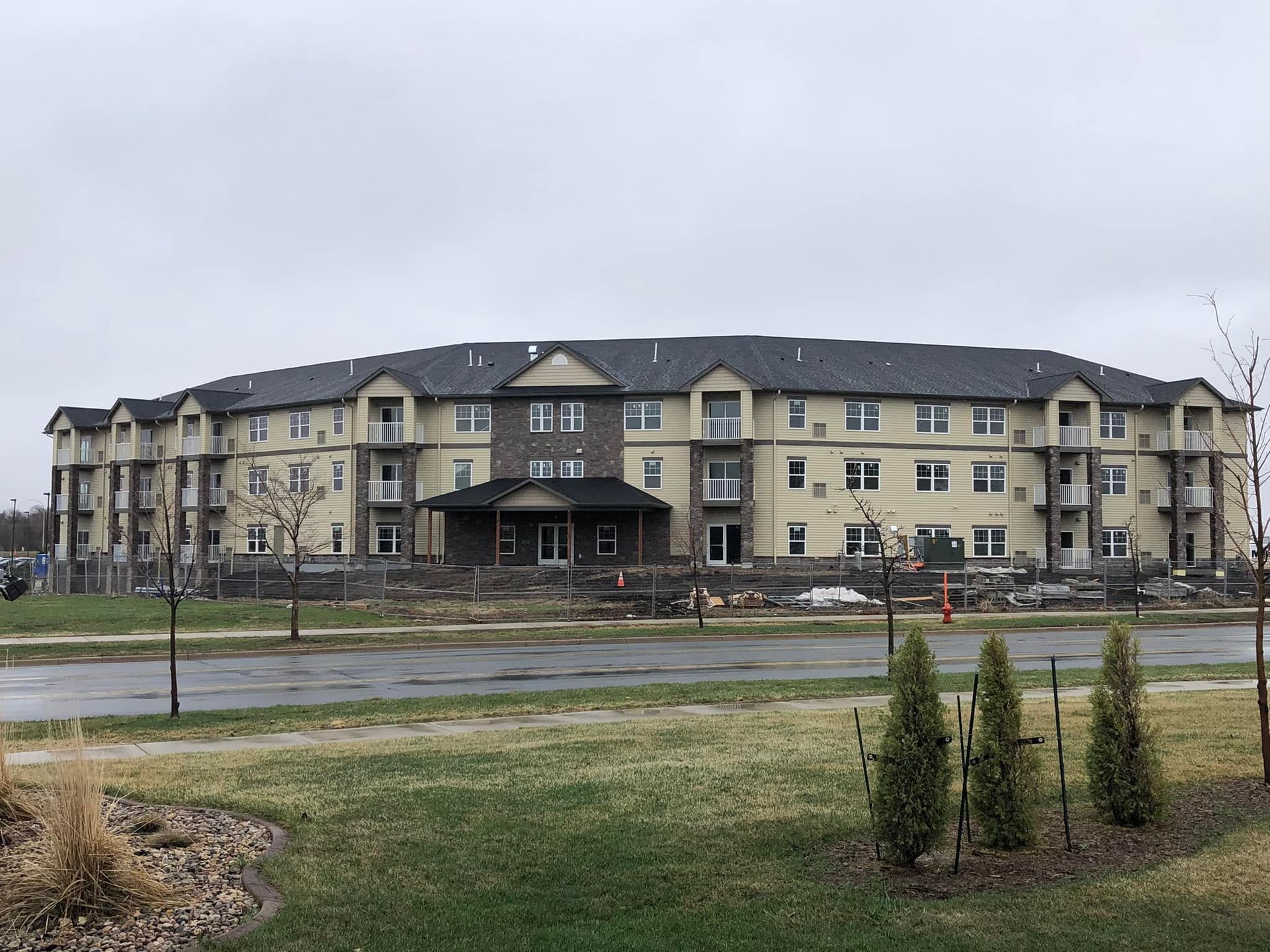 Large, multi-story apartment building with tan exterior, balconies, and dark roof under a cloudy sky.