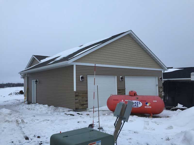 Two-car garage with tan siding and snow-covered ground, red propane tank in front.