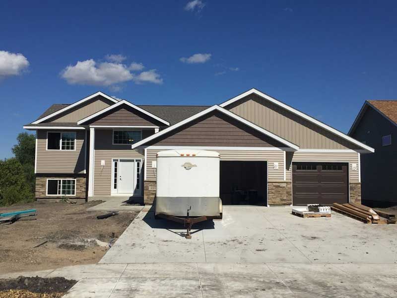 Two-story house under construction with tan siding, brown garage door, and a white trailer in driveway.