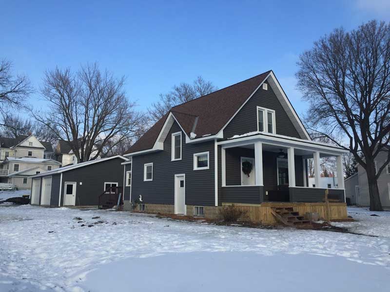 Dark gray house with brown roof, porch, and a garage on a snowy day.