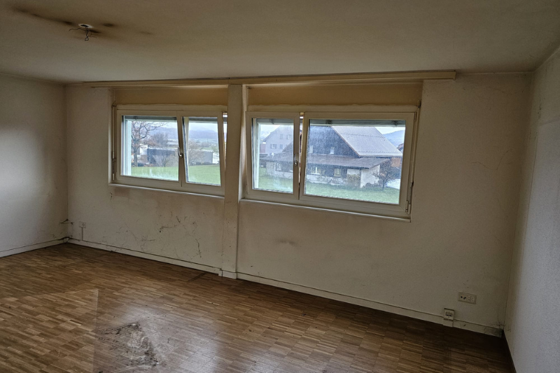 Leerer Raum mit zwei Fenstern, schmutzigen Wänden und abgenutztem Boden. Bewölkter Blick nach draußen.