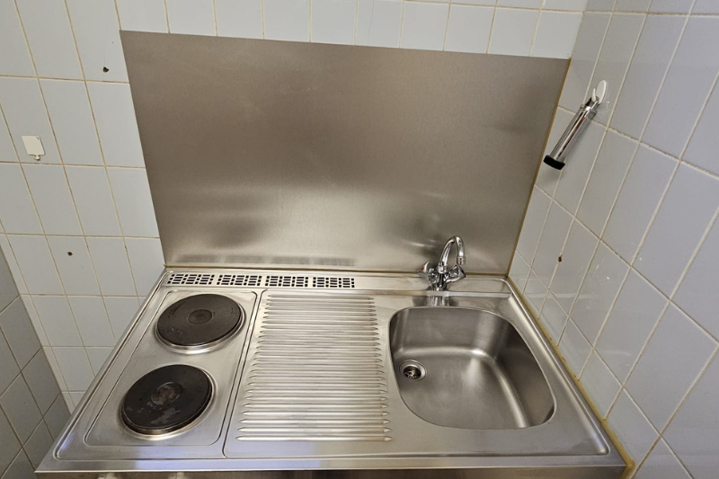 Kleine Küchenzeile aus Edelstahl: Herd mit zwei Brennern, Spüle, Spritzschutz, in weiß geflieste Wände eingelassen.