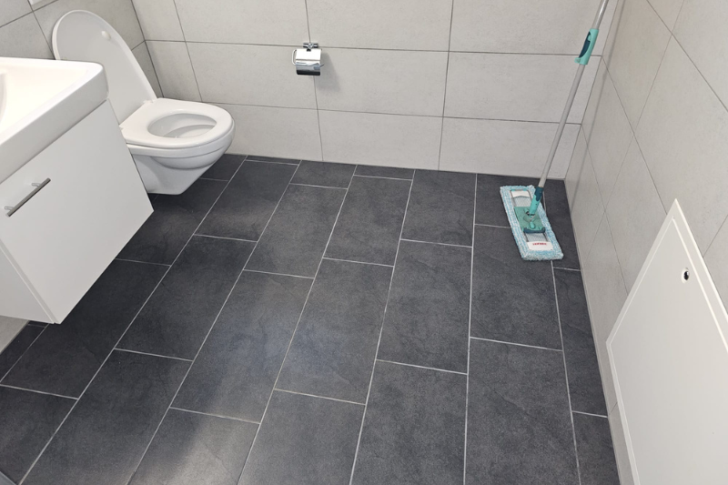 Ein Badezimmer mit grau gefliestem Boden, Toilette, Waschbeckenschrank und einem Wischmopp an der Wand.