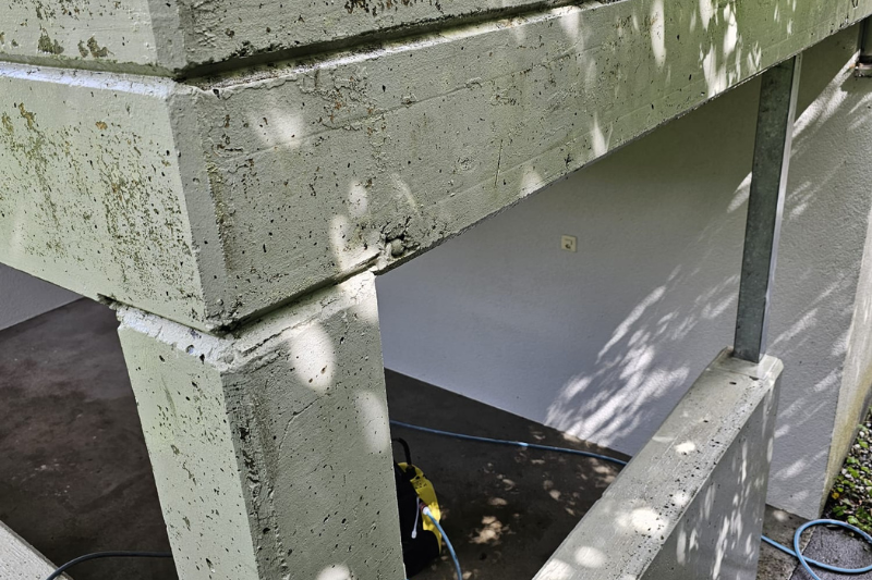 Ecke einer Betonkonstruktion mit schattigen weißen Wänden darunter; ein Metallgeländer ist sichtbar.