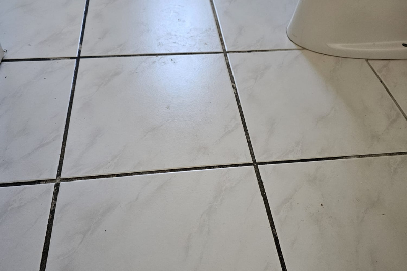 Weiß gefliester Boden mit dunklem Mörtel, teilweise sichtbar in der Nähe des Toilettensockels.