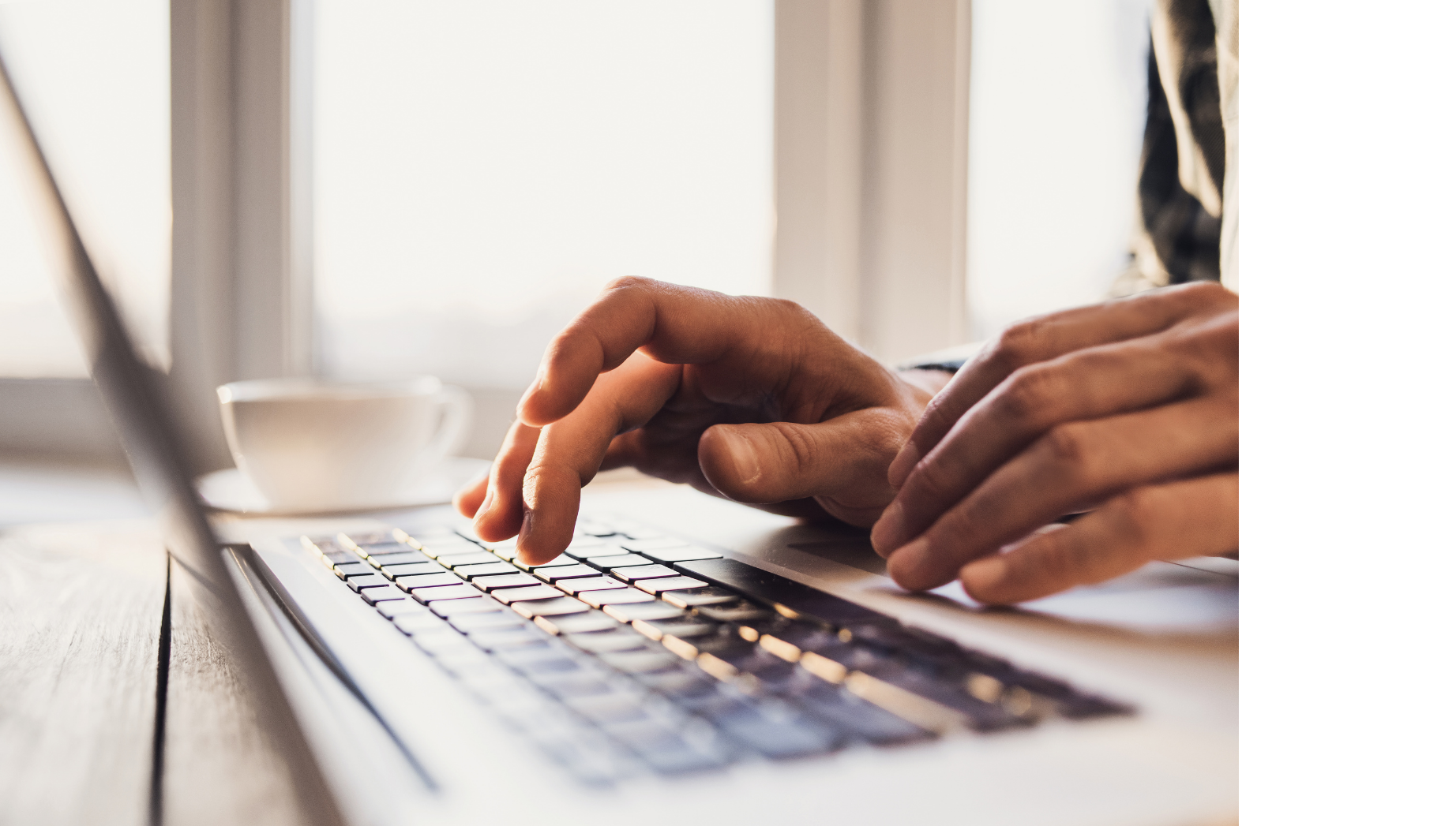 Close-up of hands typing on a laptop keyboard near a coffee cup by a bright window.