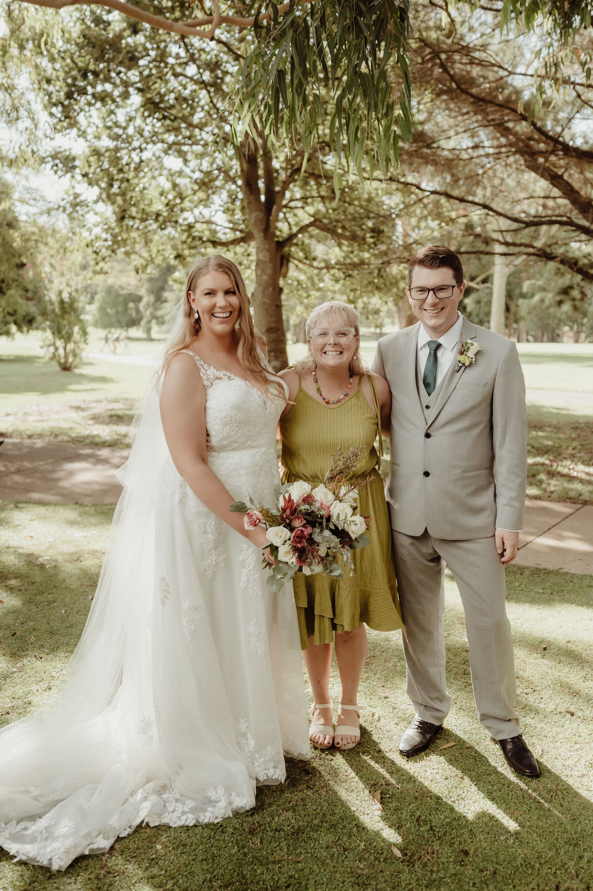 Bride and groom with a woman, smiling, in a park. Bride in white dress, groom in grey suit, woman in a green dress.