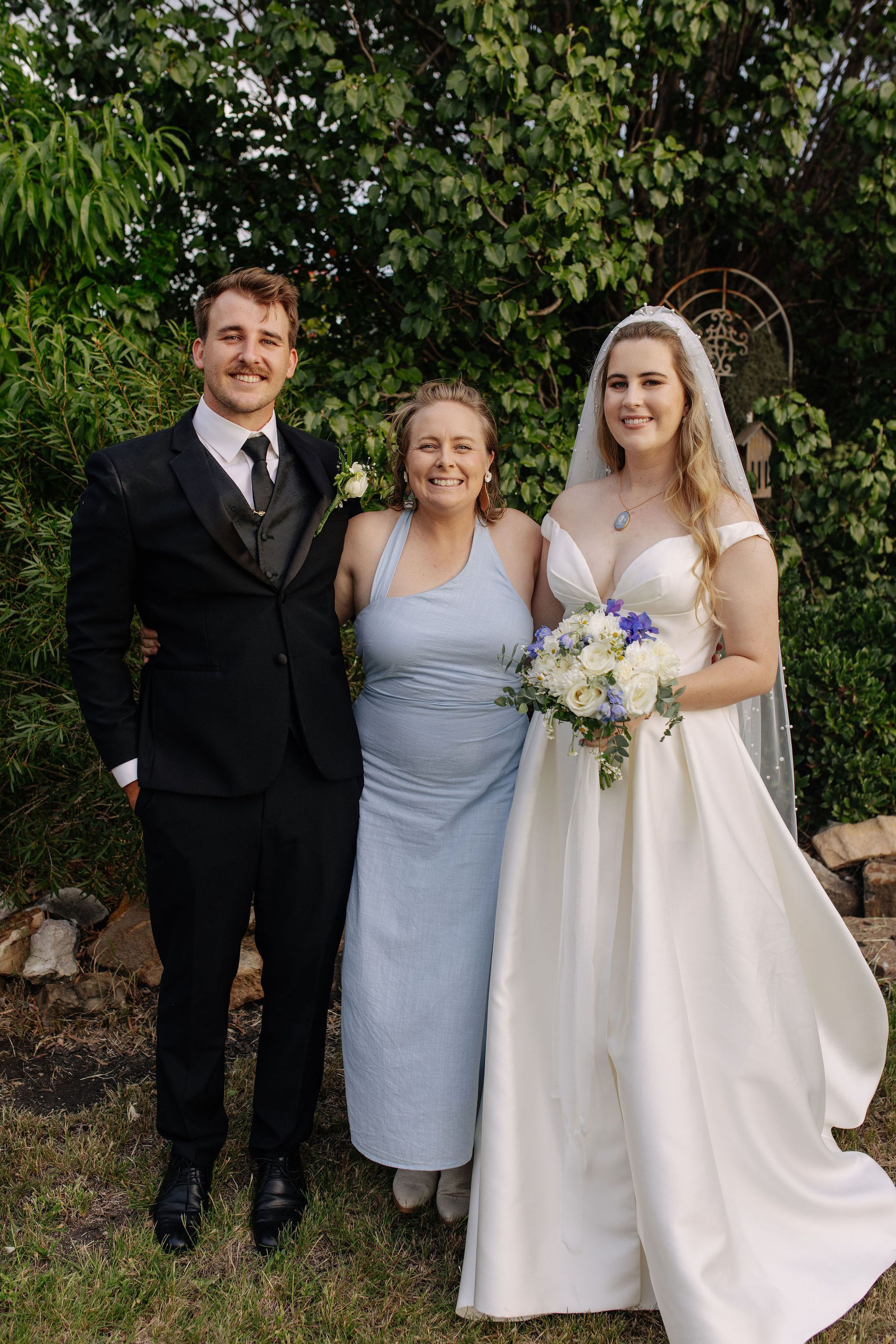 A bride in a white gown and a groom in a suit pose with a woman in a blue dress outdoors.
