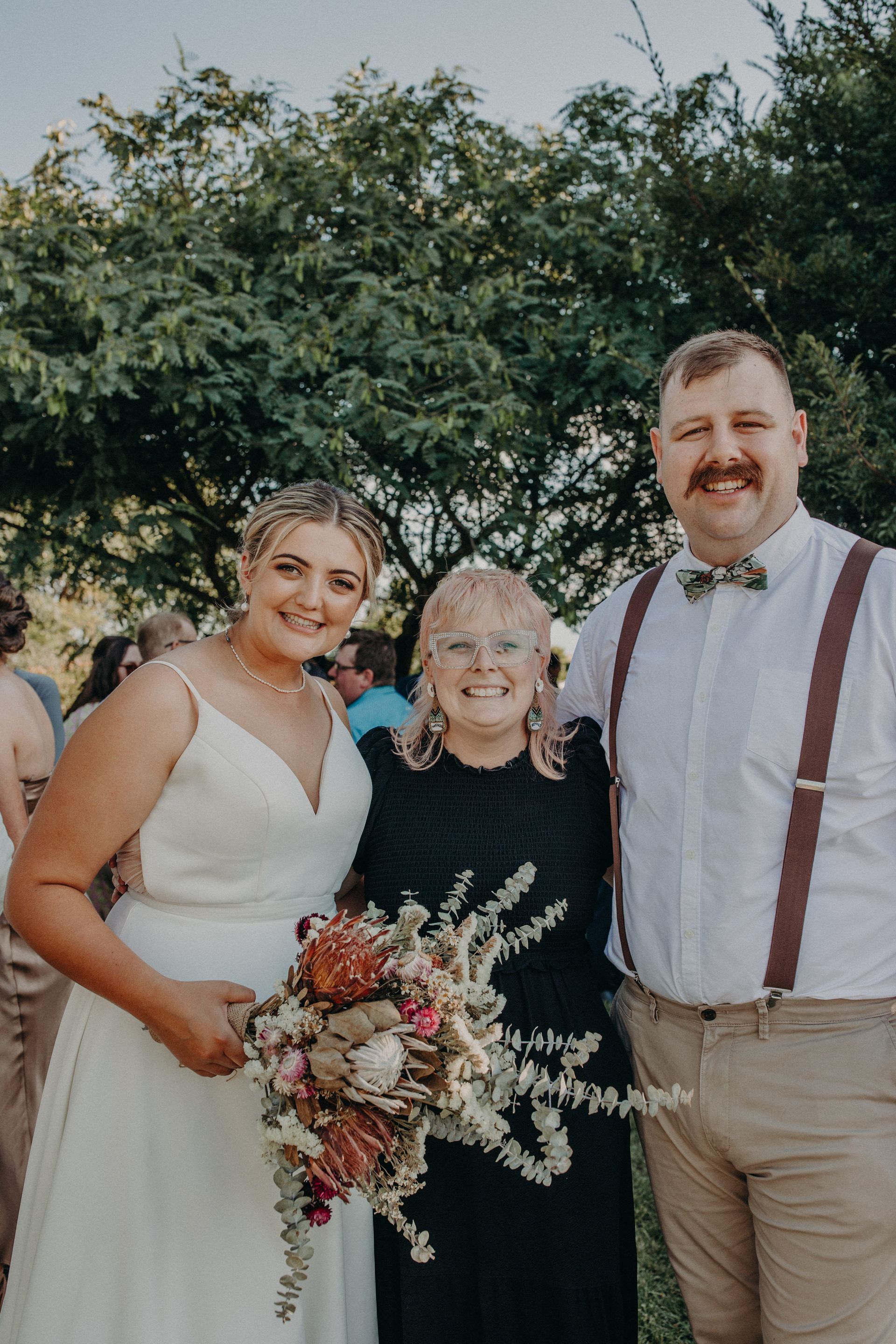 Bride and groom with a woman, posing outdoors. Bride holds bouquet, all smiling.