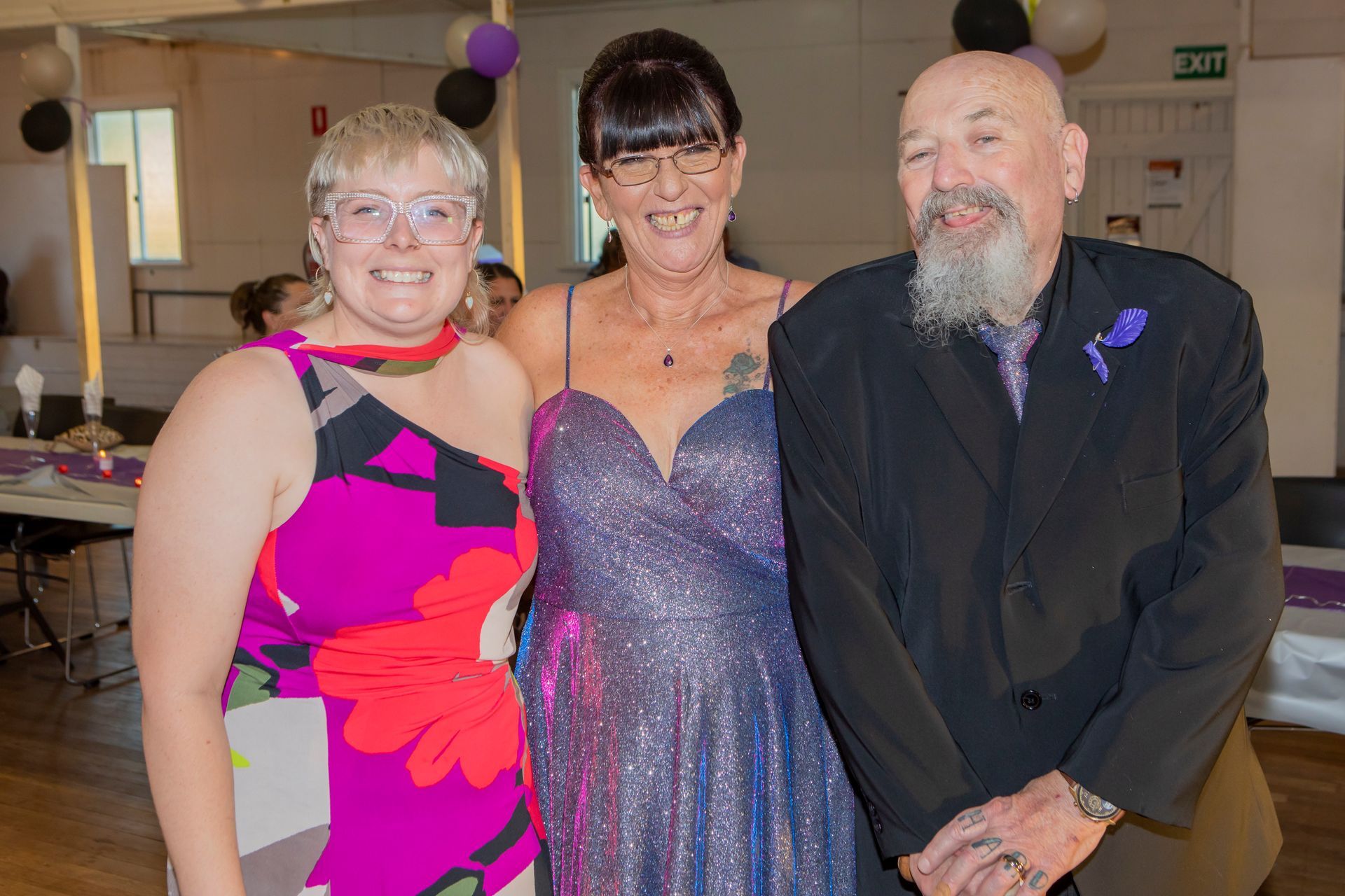 Three people smiling together at an event. Woman in sparkly blue dress, another in floral pink top, man in suit.