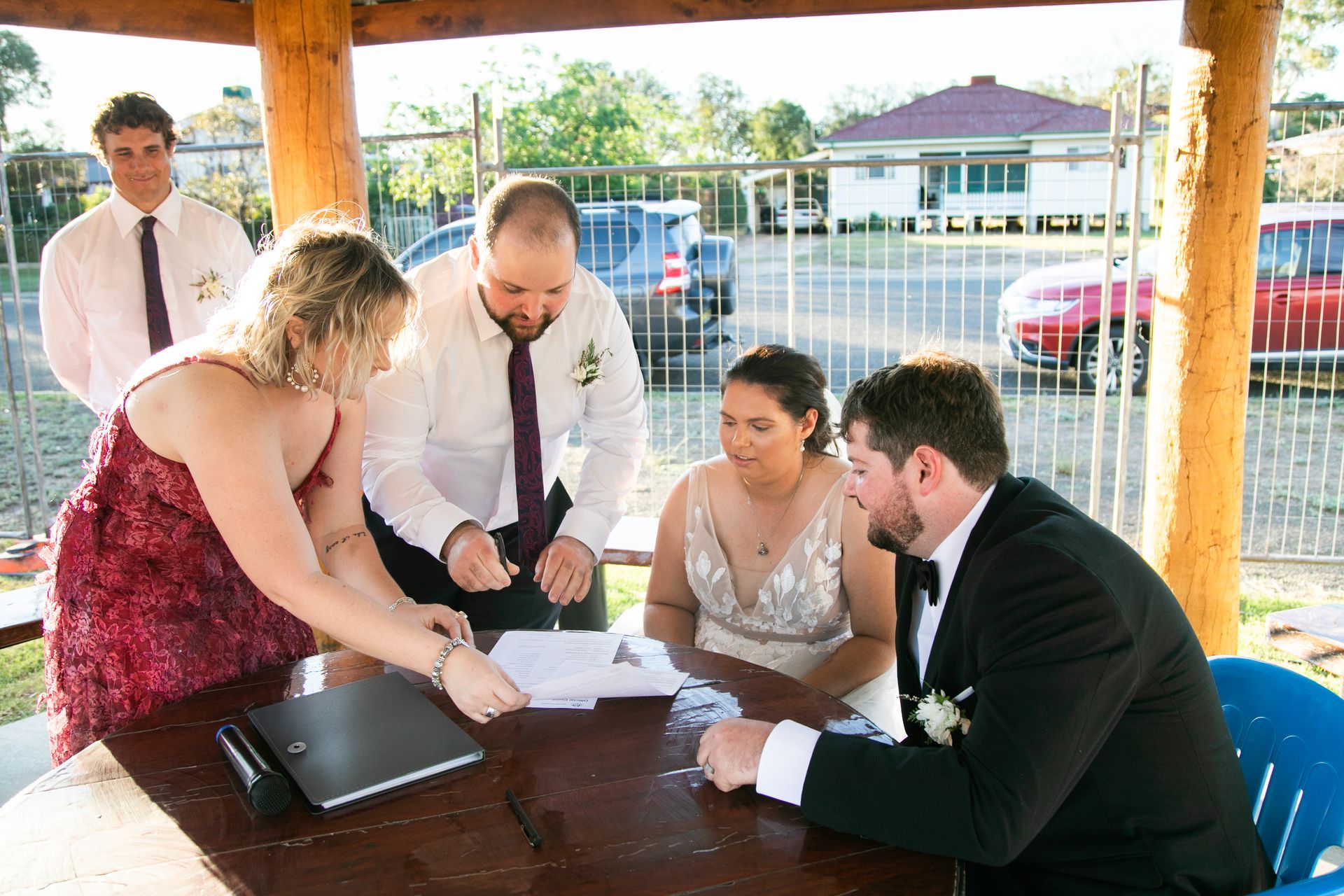 Wedding ceremony: couple signing documents at a table, officiant and witness present, outdoor setting.