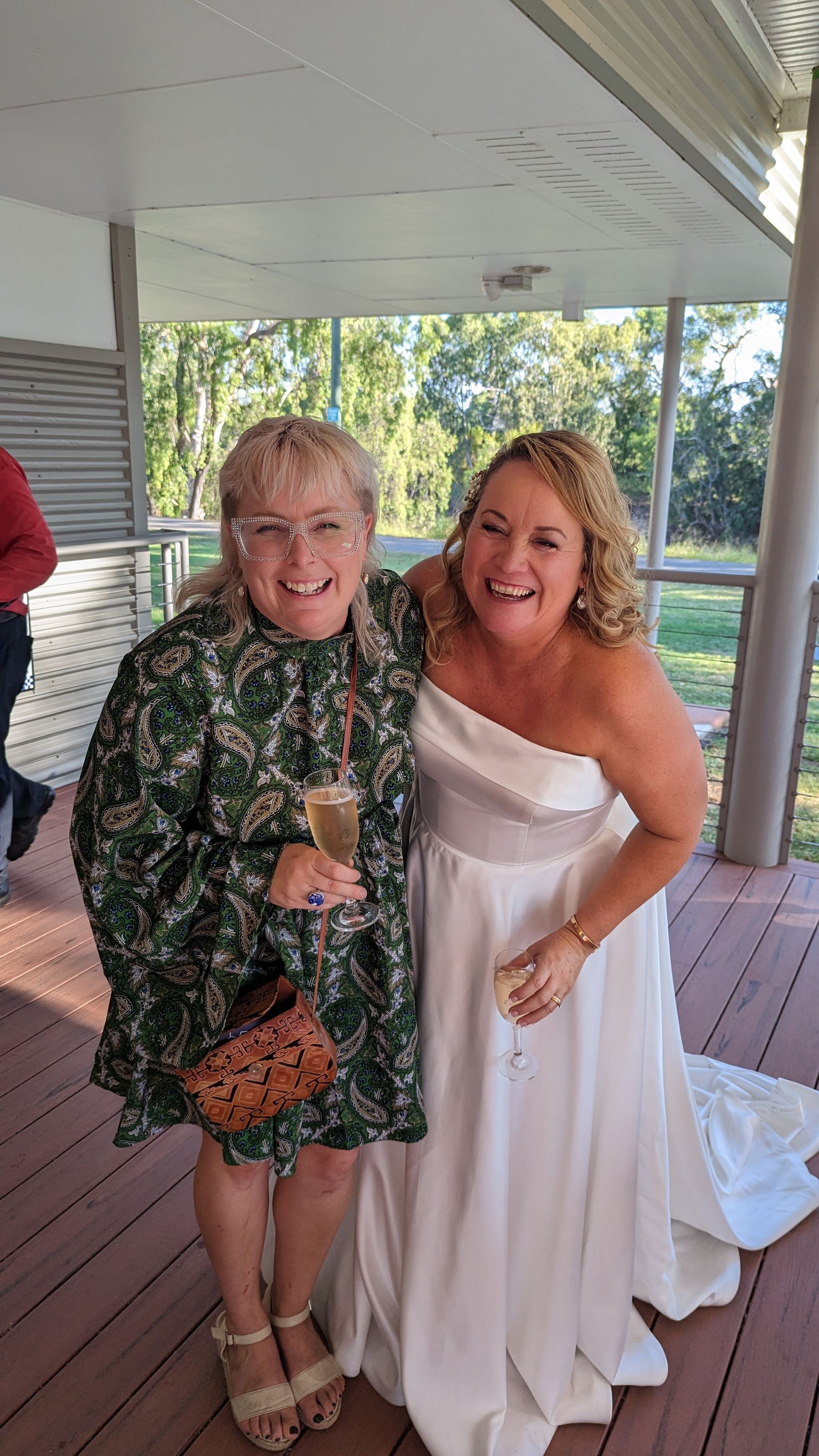 Two smiling women, one in a floral dress holding a glass, the other in a white strapless wedding gown, outdoors.
