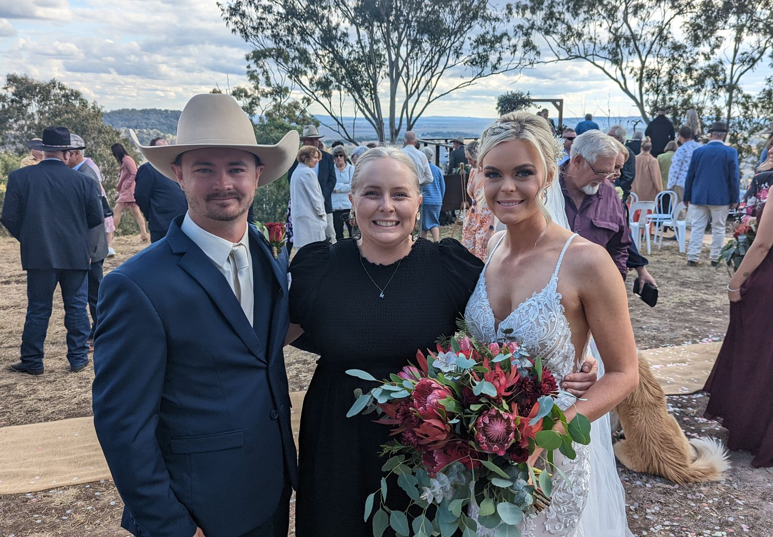 Bride and groom with officiant at outdoor wedding. Bride holds bouquet, groom in cowboy hat. Guests in background.