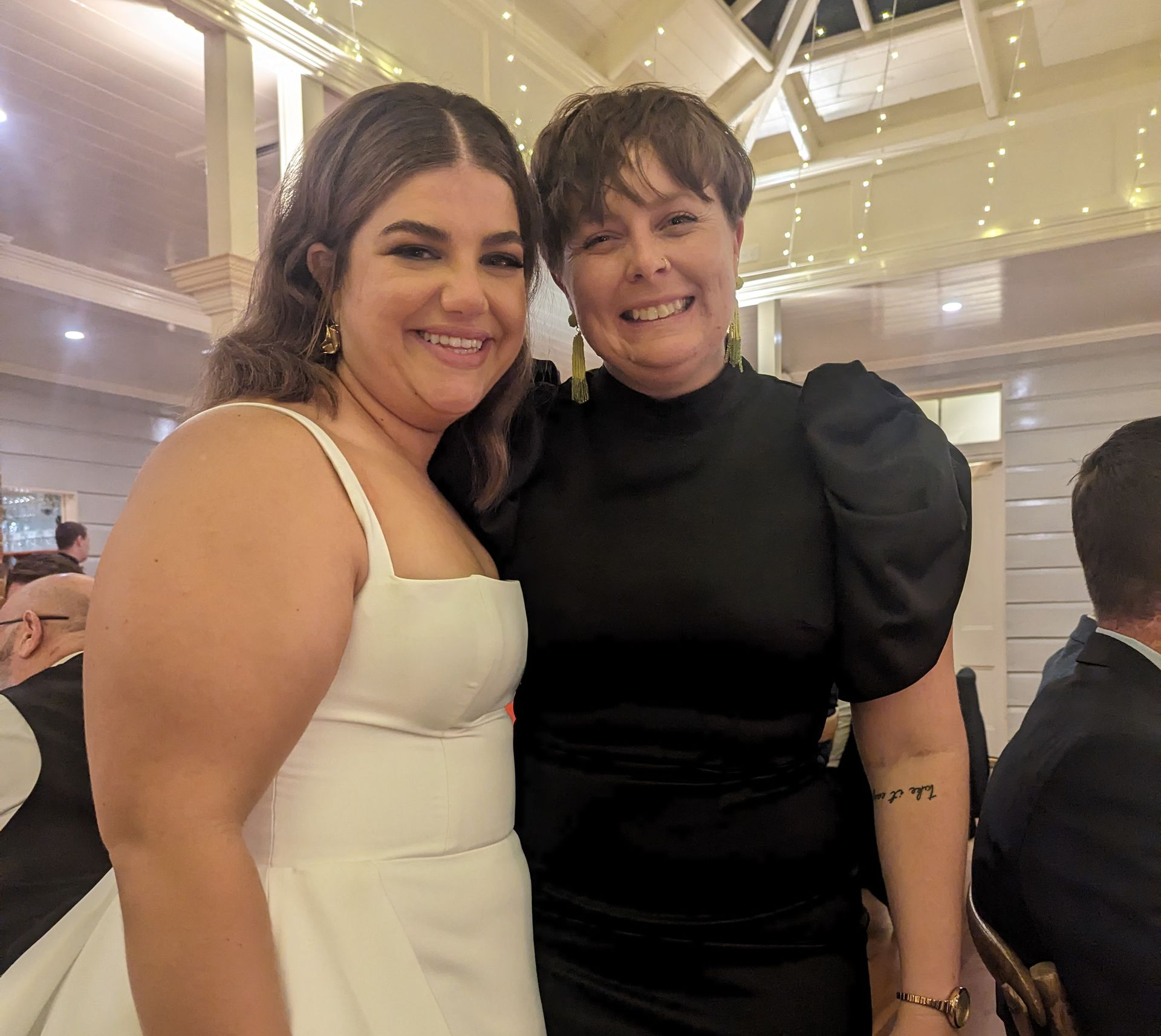Two smiling women pose indoors, one in a white dress, the other in black, both with arms around each other.